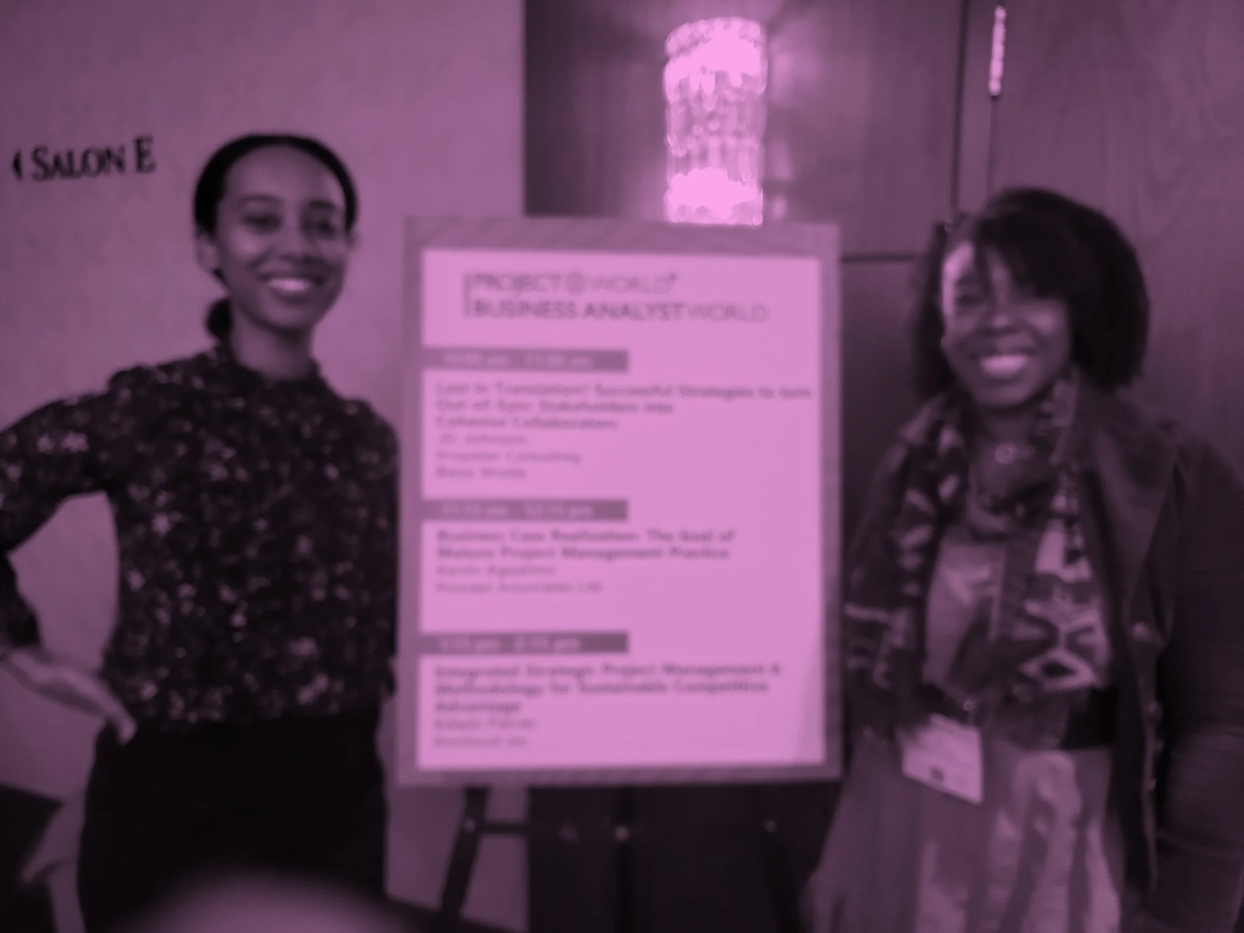 Two women standing in front of a poster at a conference or event. The woman on the left has dark hair tied back and is wearing a patterned blouse, smiling. The woman on the right has curly hair and is also smiling. The poster between them is titled 'Project Outreach' and discusses content related to business and management.