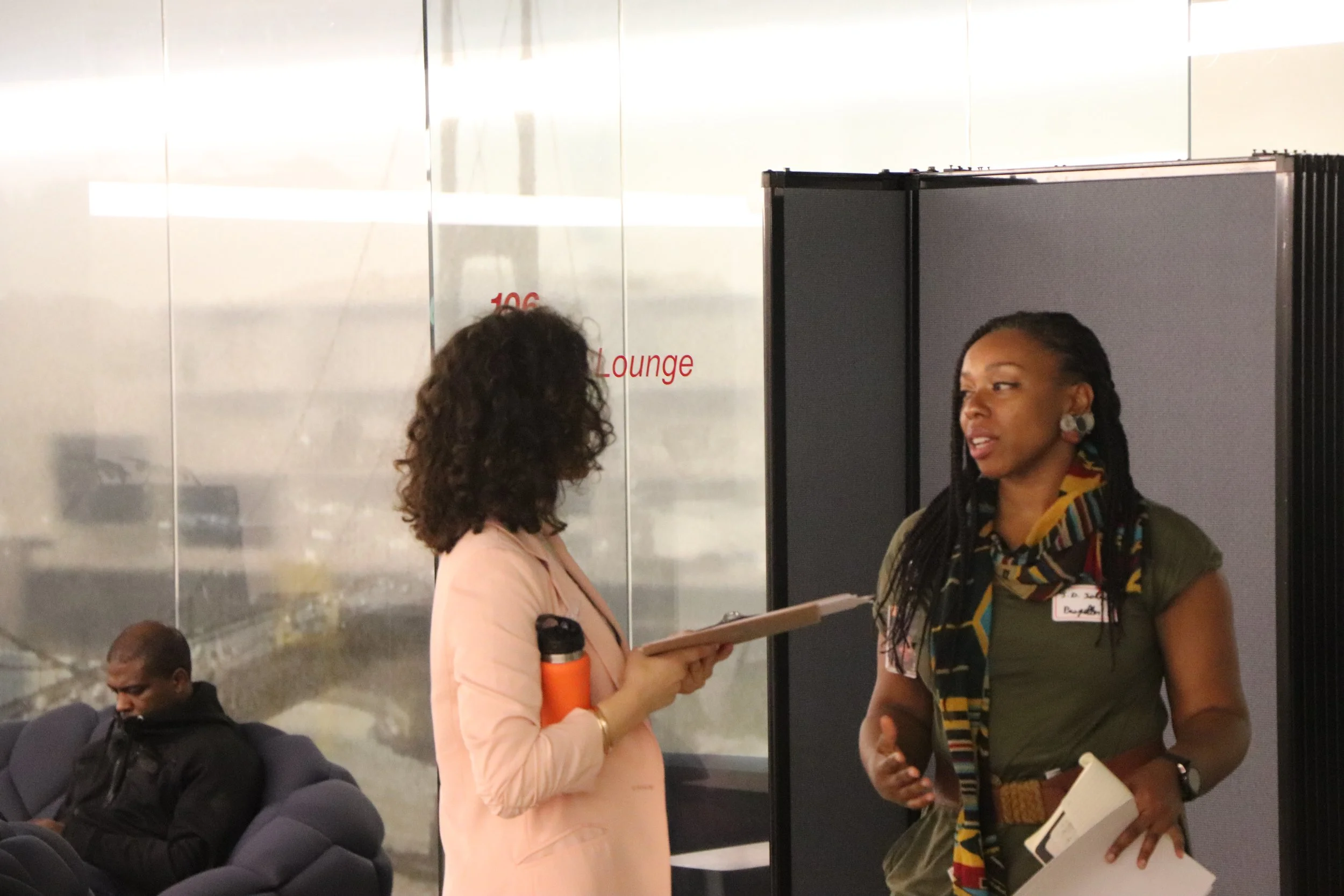Two women are having a conversation in an airport or similar setting. One woman is holding a clipboard and a water bottle, while the other woman, possibly an employee with a name tag, is listening. There are airport seats and other people in the background.