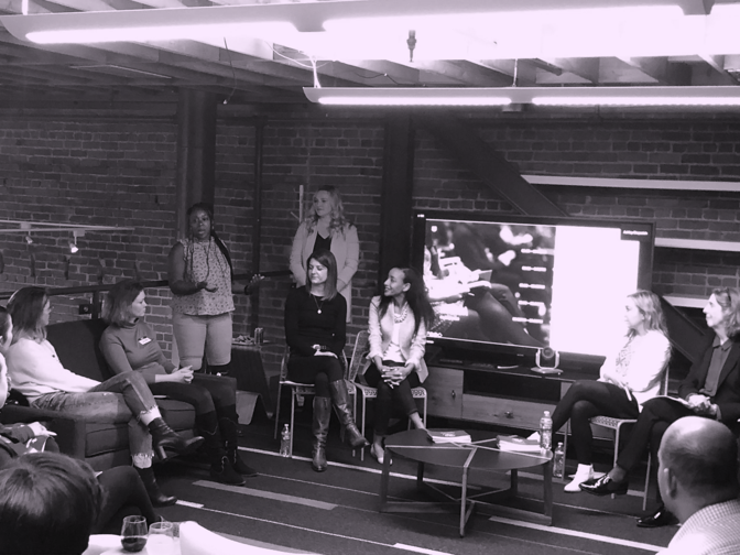 Group of women participating in a panel discussion in a room with exposed brick walls, with some seated on chairs and one standing, in front of a television screen.