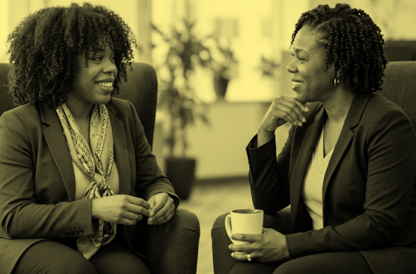 Two women sitting and talking in an office, with one holding a cup of coffee.