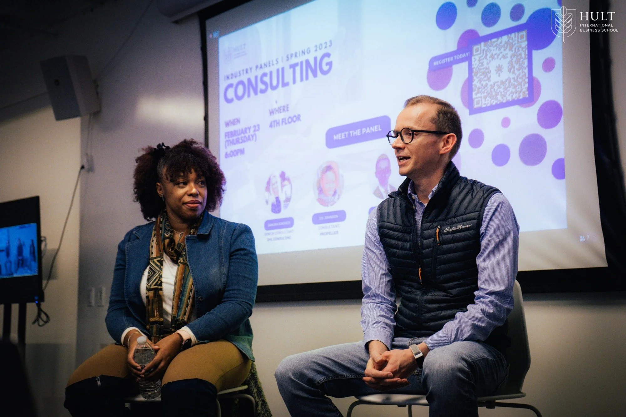 A woman and a man are seated in front of a presentation screen during a panel discussion. The screen displays information about an industry panel event at Hult International Business School, scheduled for February 23rd at 4 PM, with a QR code for registration. The woman has curly hair and is wearing a denim jacket, while the man is wearing glasses and a black quilted vest over a shirt.