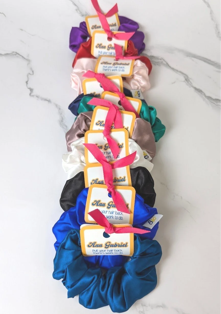 A colorful display of hair scrunchies arranged vertically with attached tags reading "Ann Gabriel" and a message to pull the hair back for work to do, on a white marble surface.