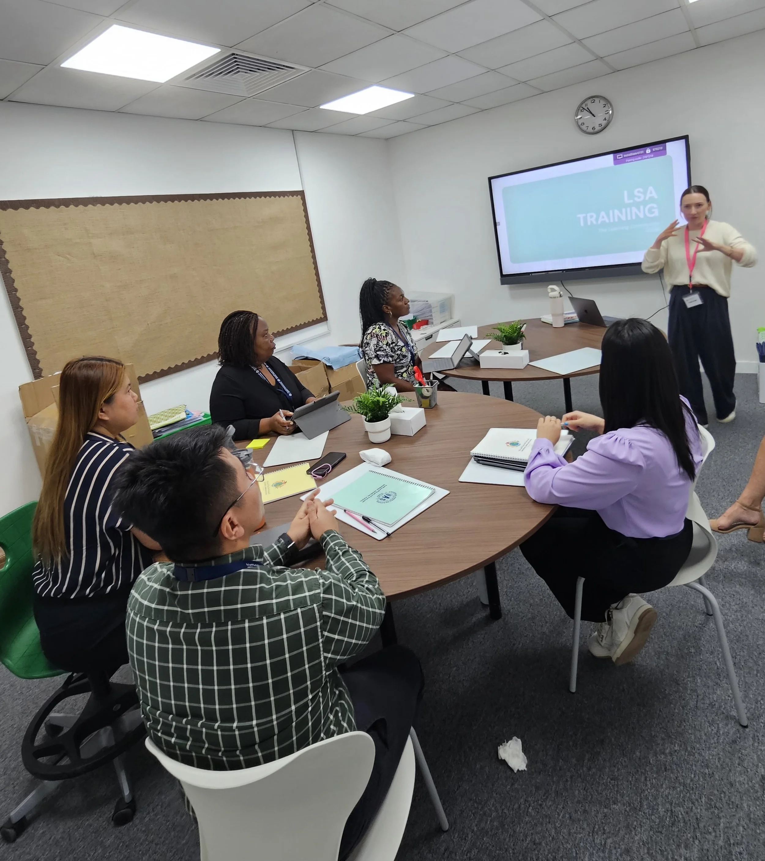 A group of six people attending a training session in a conference room, with a woman presenter gesturing in front of a large screen displaying 'LSA TRAINING' and a clock on the wall.