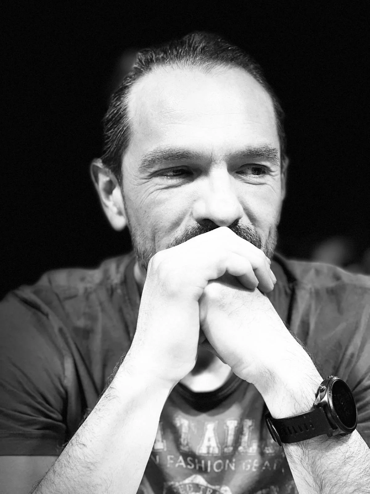 Black and white photo of a man with a beard and mustache, resting his chin on his clasped hands, wearing a watch, with a contemplative expression.