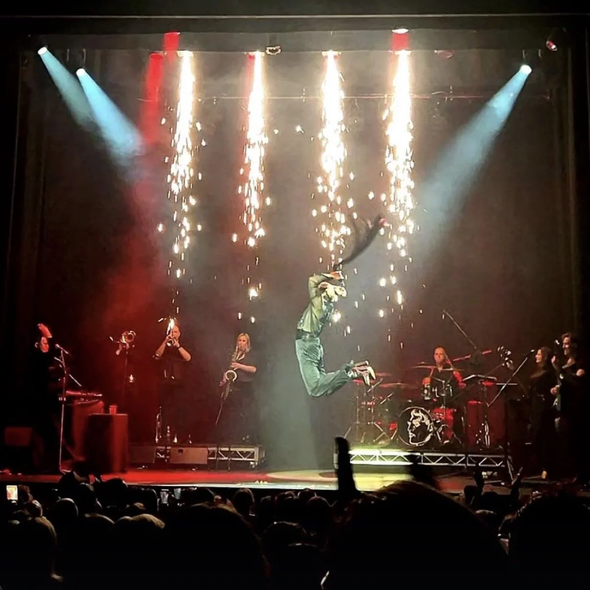 Reuben Kaye 'Hard to Swallow' at the Melbourne International Comedy Festival 2026. Performer jumping on stage during a concert with sparklers and laser lights, with band members playing instruments and an audience watching.