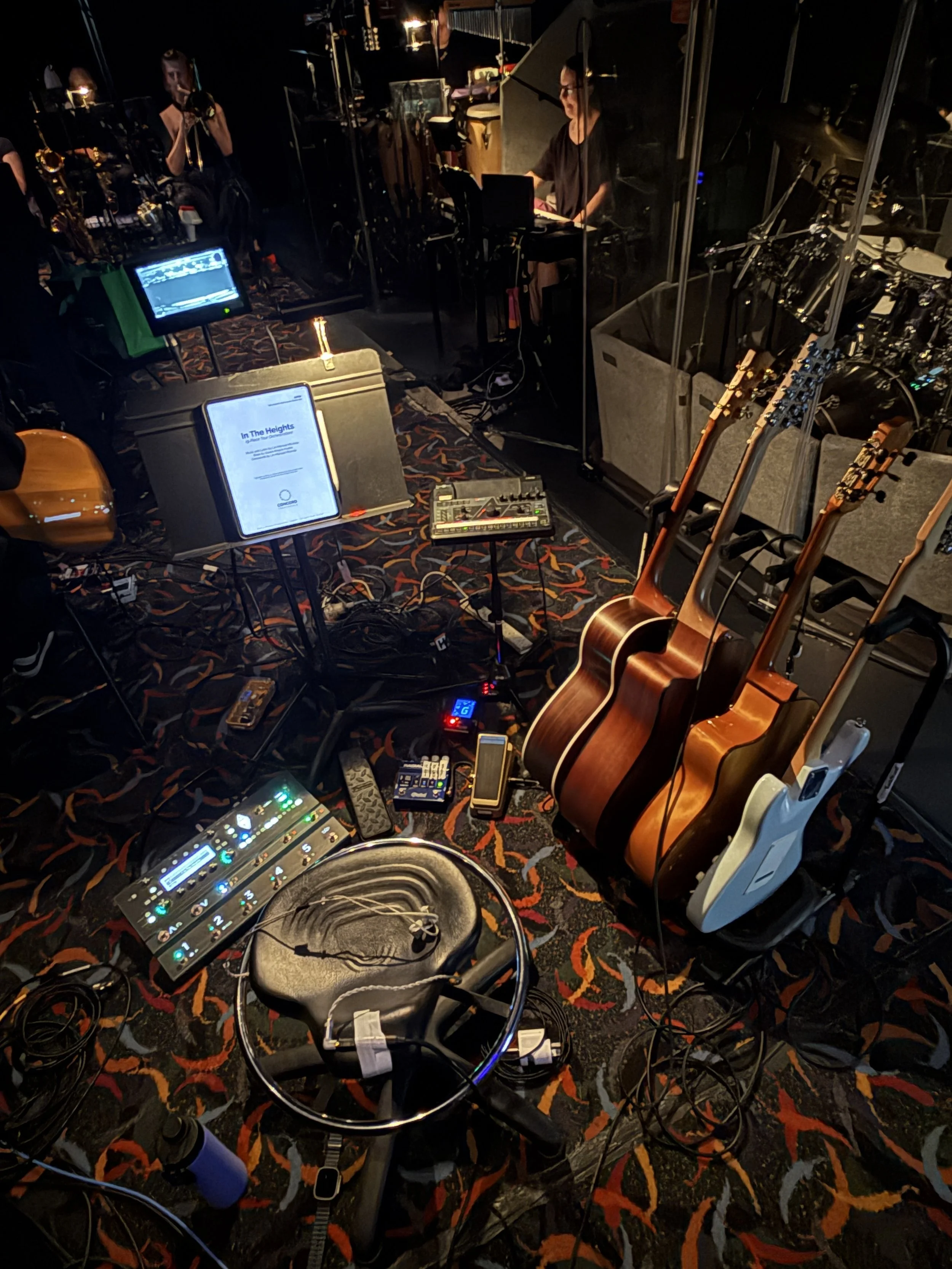 A recording studio with various musical instruments, including guitars, and electronic equipment. There are people working with microphones and mixing consoles in a dark, cozy environment.
