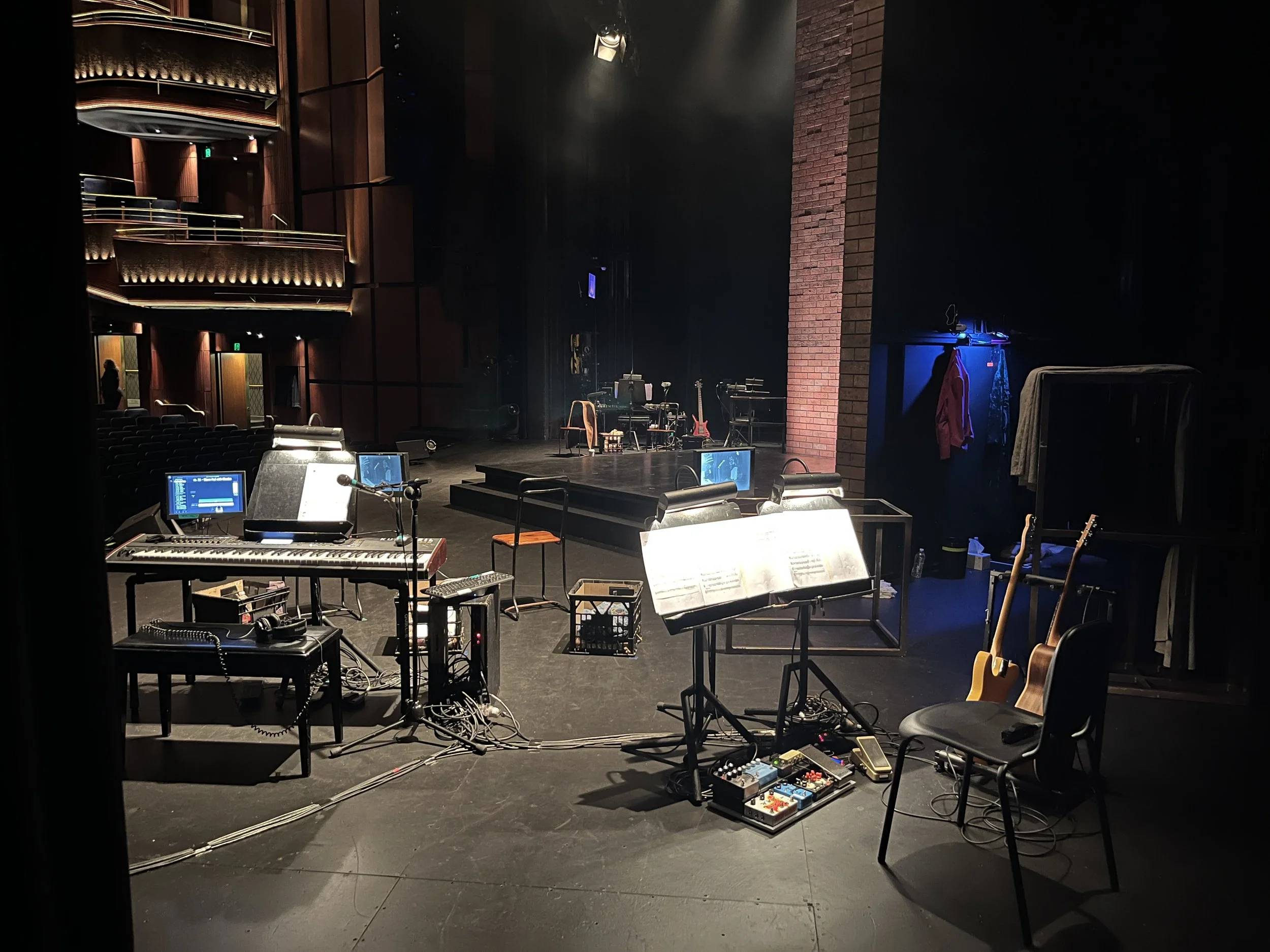 Empty stage with musical instruments including guitars, keyboard, and pedals, music stands, chairs, and various audio equipment set up, in a theatre or concert hall with seating balconies.
