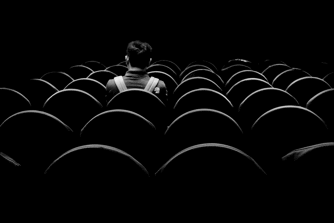 A person sitting alone in an empty theater with rows of empty seats, seen from behind in black and white.