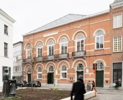 Historic red brick building with arched windows and balconies on a city street, with pedestrians and parked cars.