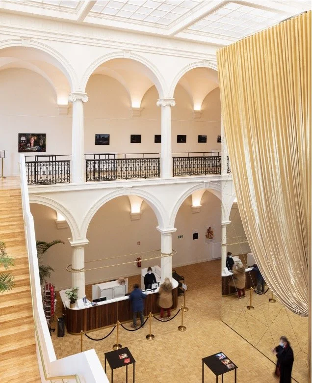 Interior of a museum or gallery with white arches, a mezzanine level, framed artworks on the walls, a reception or ticket counter with people, and a large golden curtain.
