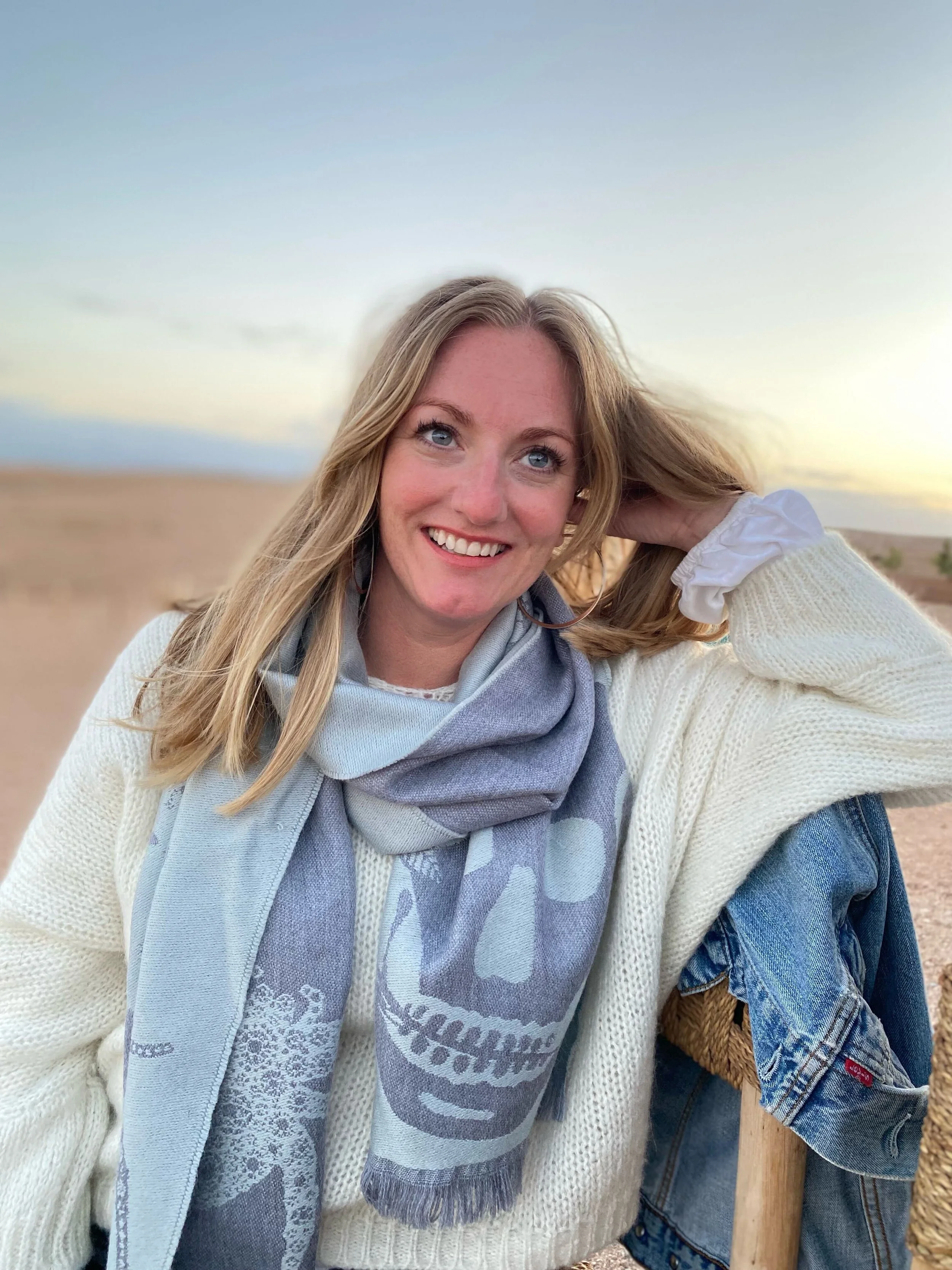 A woman with long blonde hair smiling outdoors at sunset, wearing a cream sweater, a skull-themed scarf, and a white shirt with a denim jacket draped over her arm.