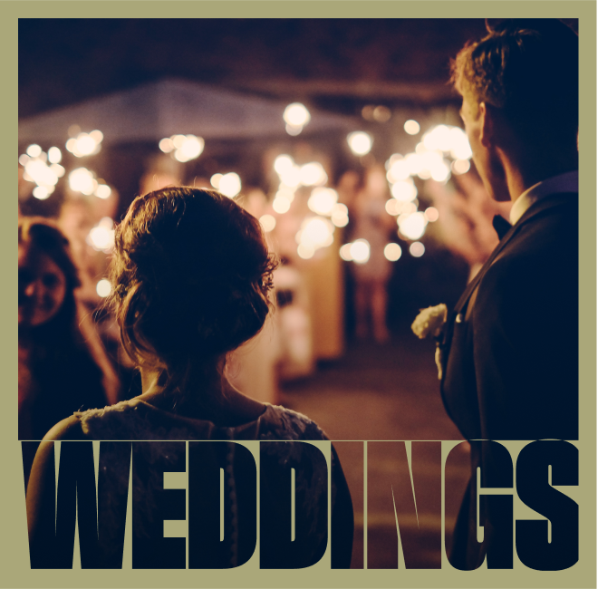 A couple at a wedding reception, with sparklers and guests in the background, during nighttime celebration.