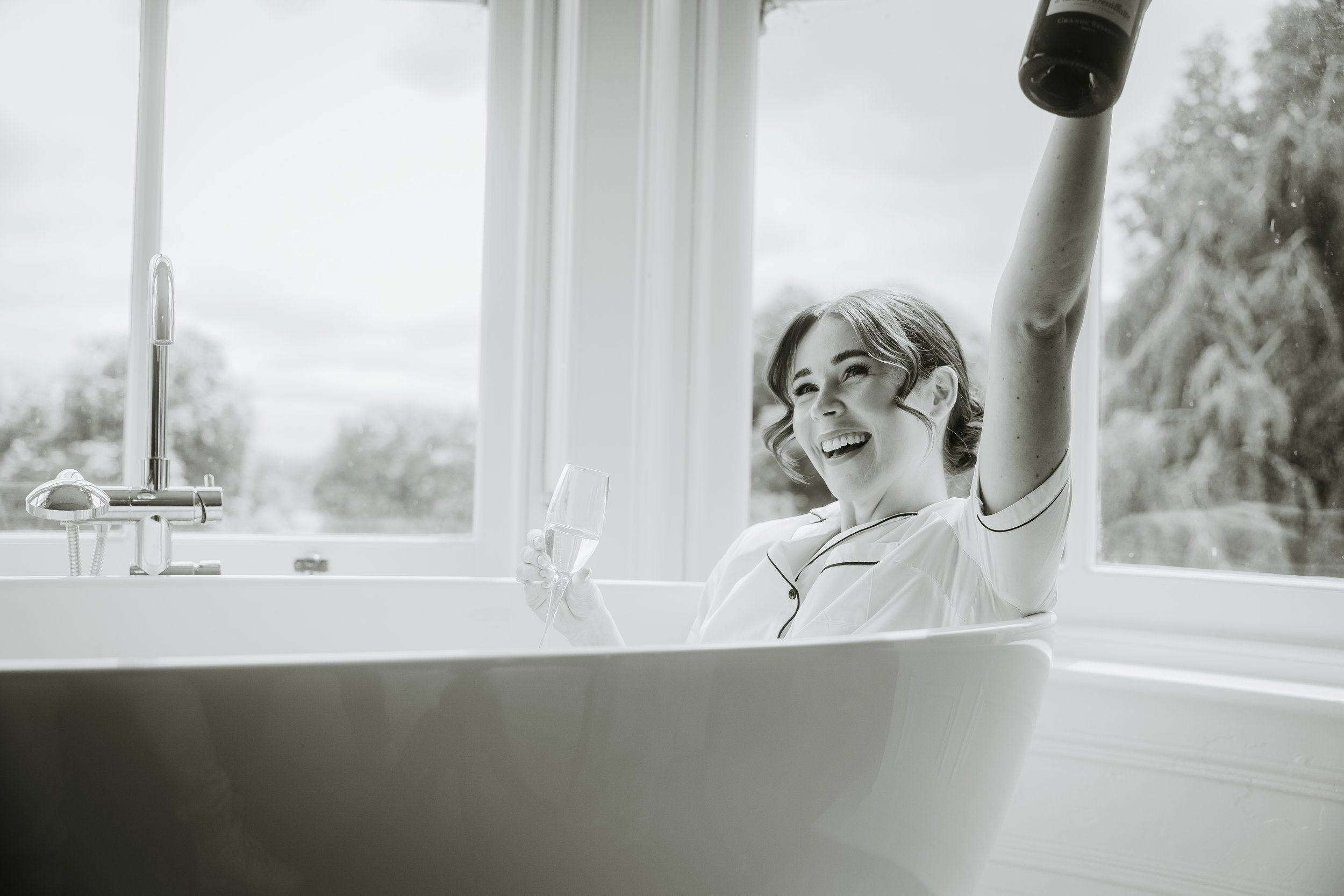 Woman in a bathtub wearing a pajama top, holding a wine glass and raising a wine bottle, looking happy, with large windows and trees visible outside.