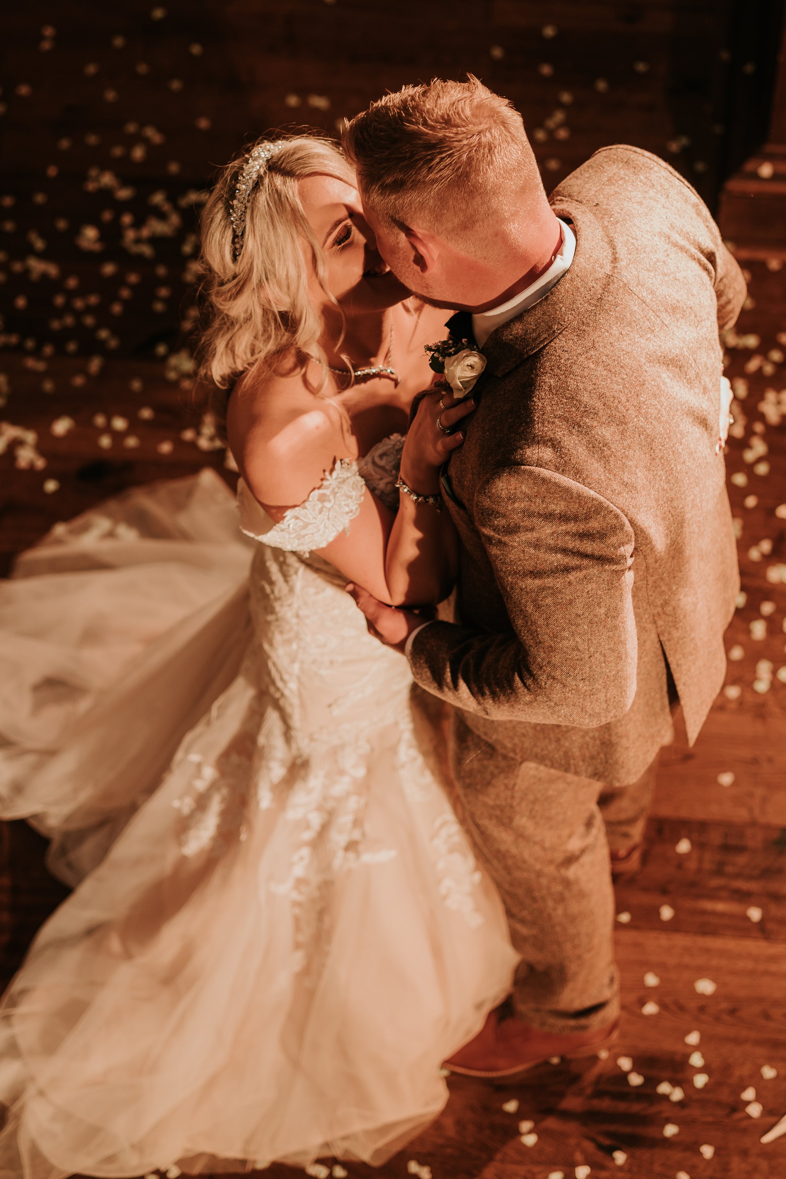 Bride and groom kissing and dancing on a wooden floor surrounded by flower petals.