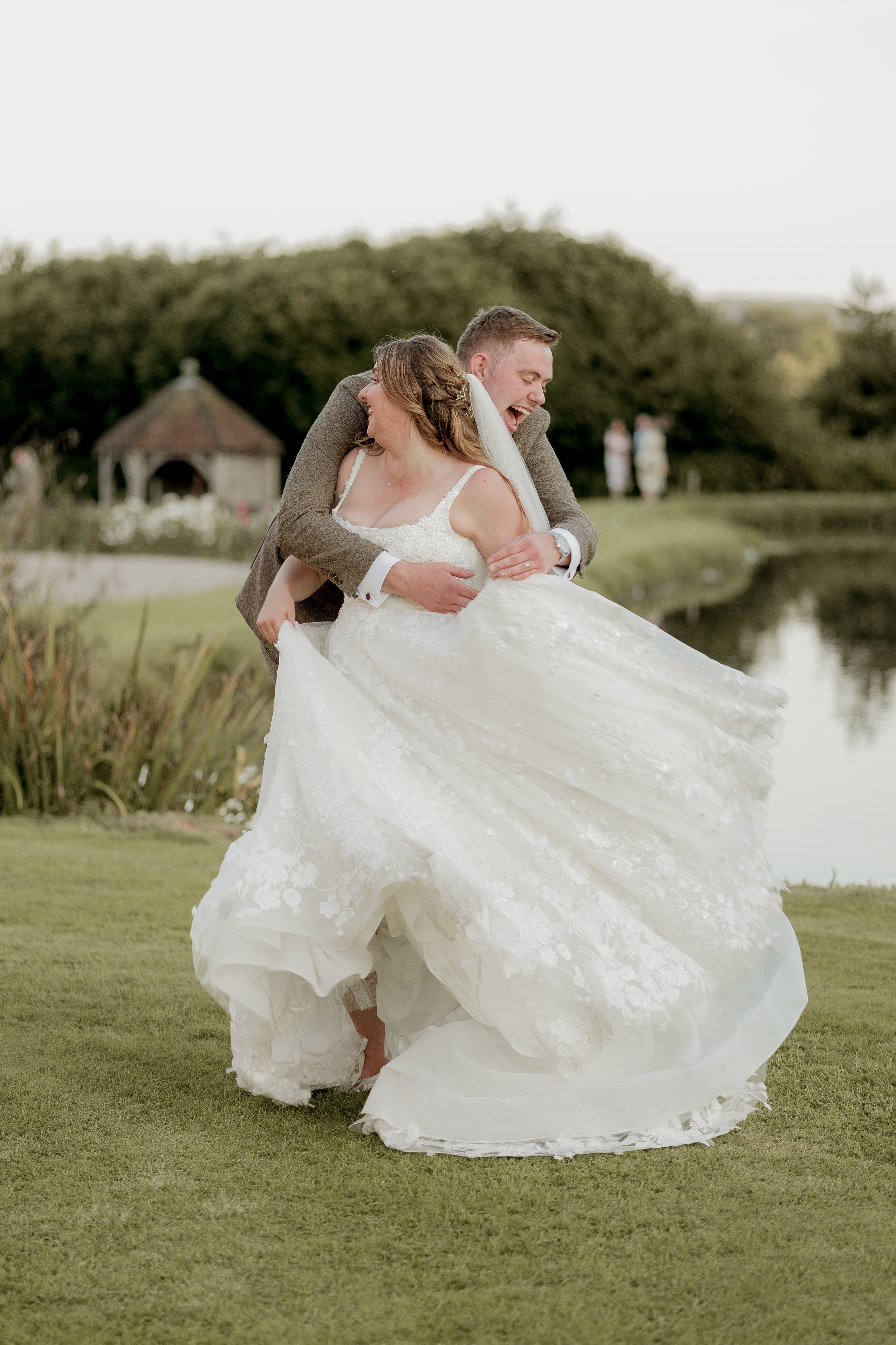 Bride and groom laughing and spinning joyfully on a grassy lawn by a pond, wearing a white wedding dress and a suit.