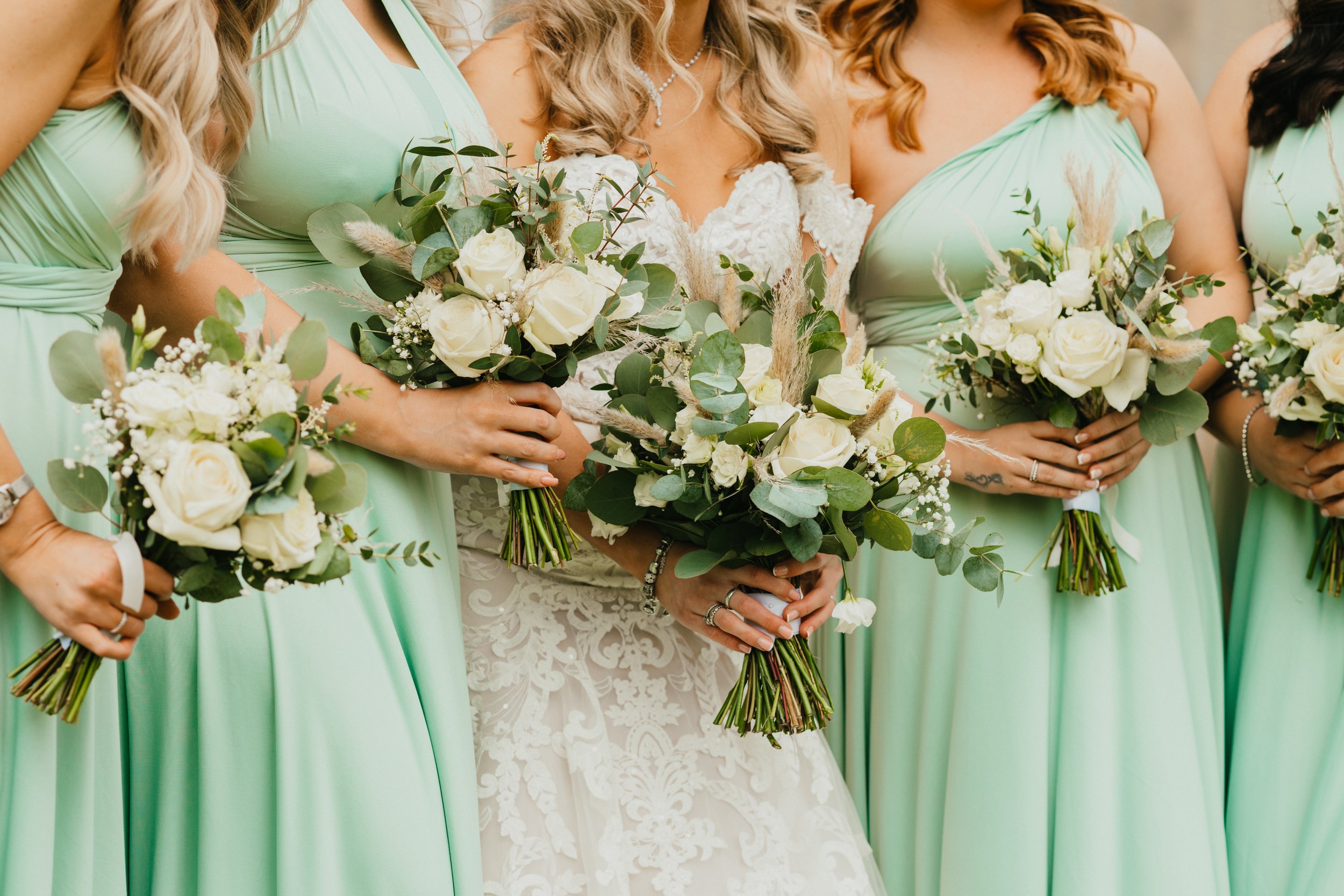 Bride in lace dress with bridesmaids in green dresses holding white flower bouquets.