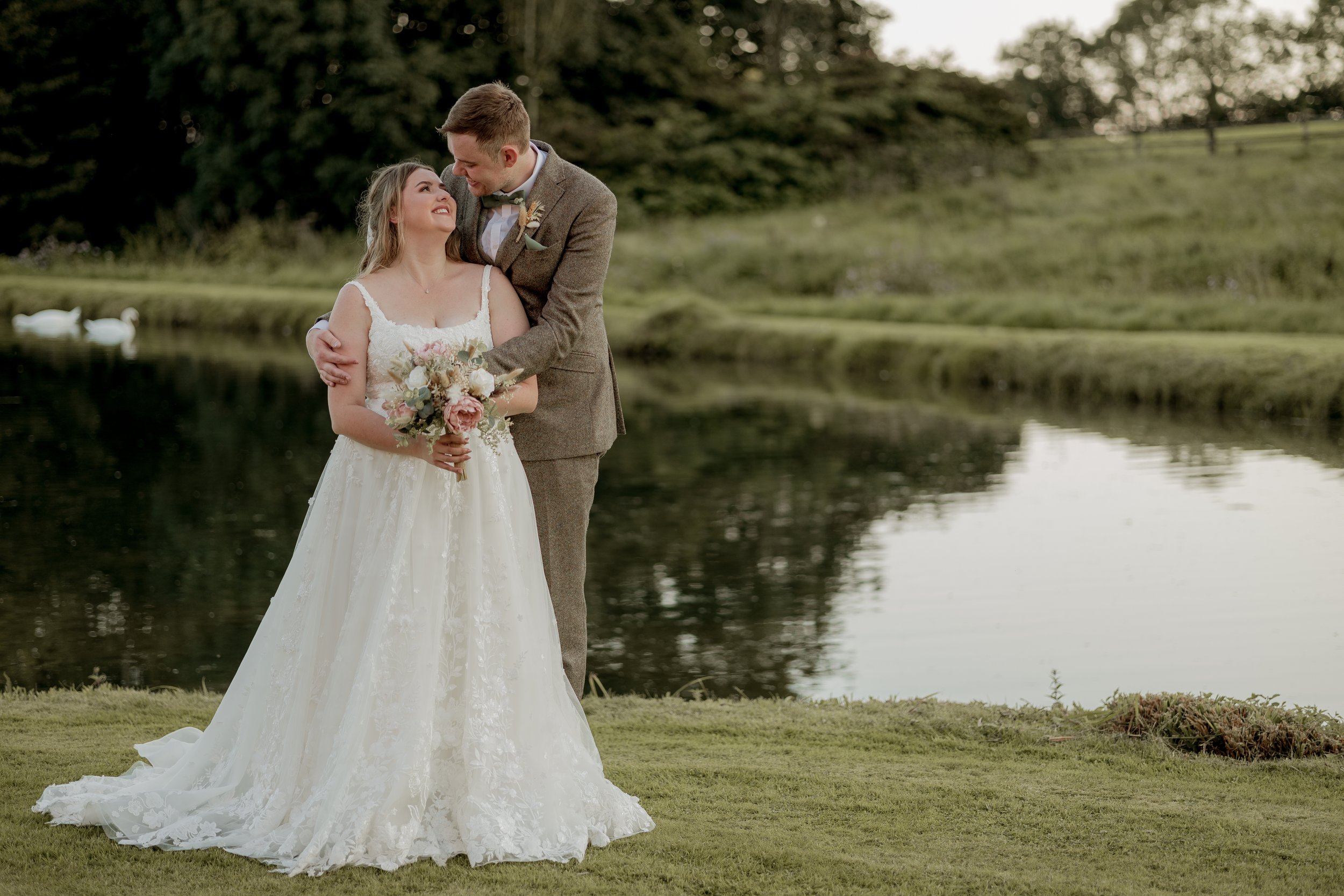 A bride and groom stand lovingly by a calm pond, surrounded by greenery. The bride is in a white lace dress, holding a bouquet, while the groom in a suit embraces her. Swans are visible on the pond.