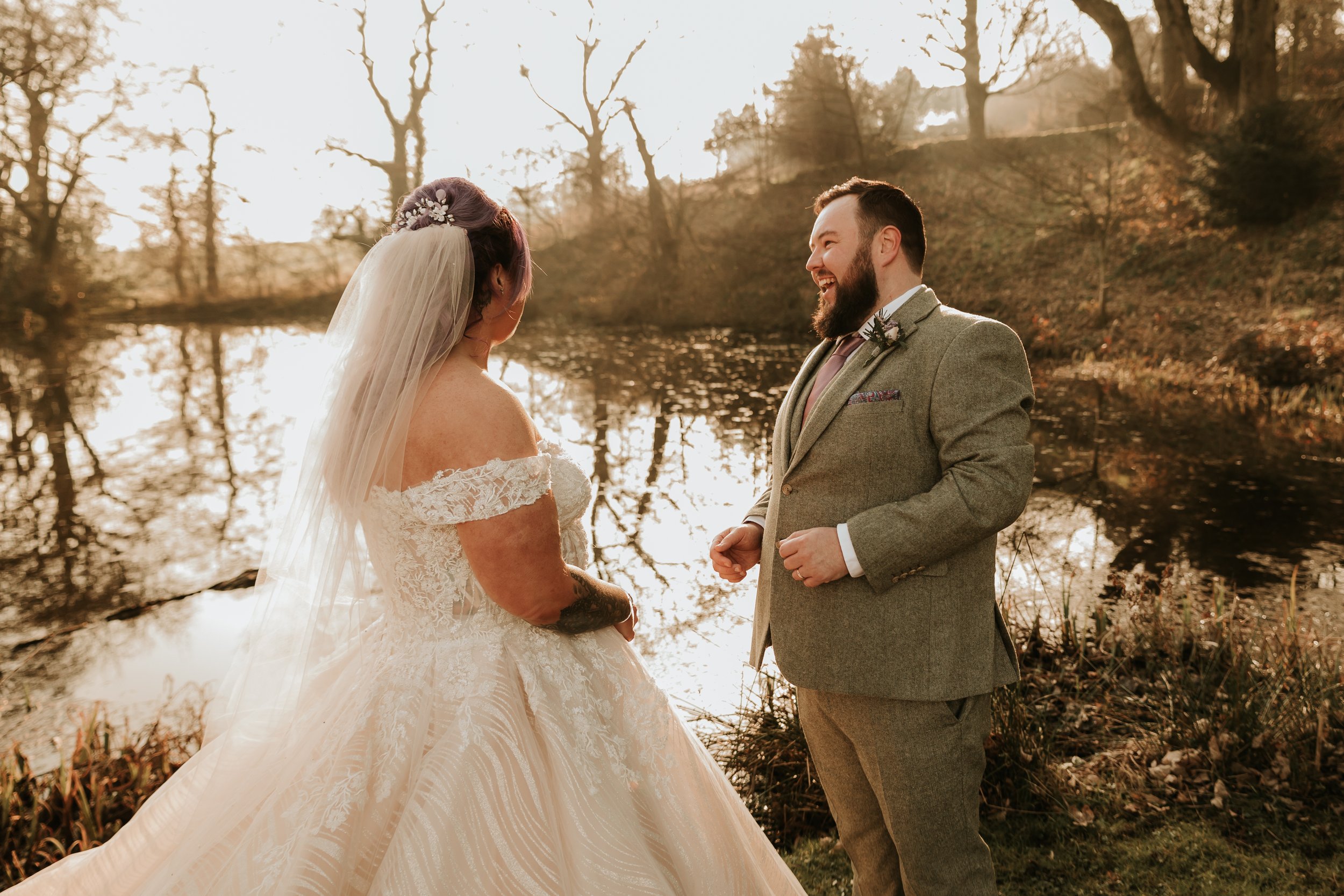 A bride and groom smiling at each other by a pond, surrounded by trees in a park setting.