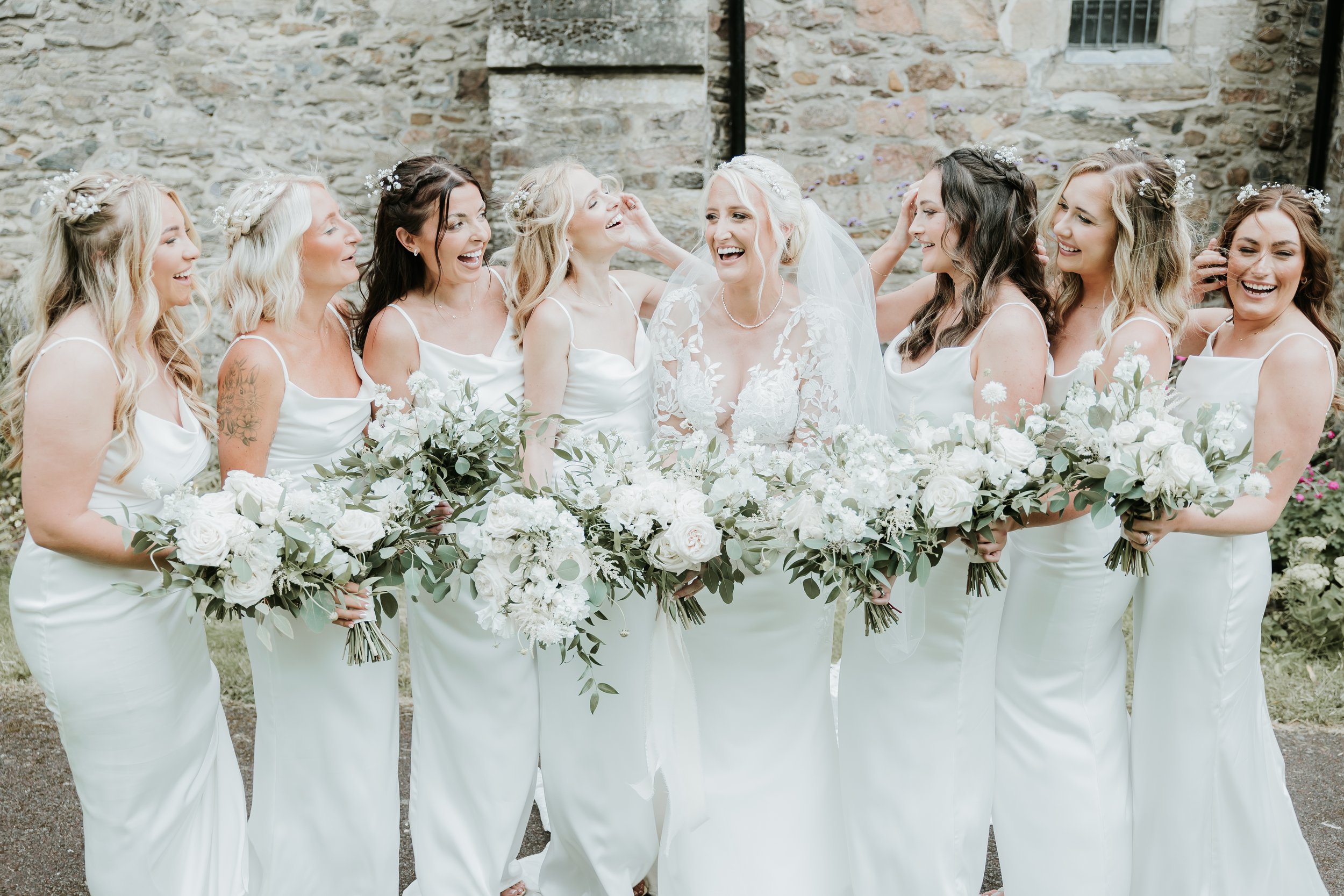 Bride and bridesmaids wearing white dresses, holding bouquets of white flowers, standing together outside, smiling and laughing.