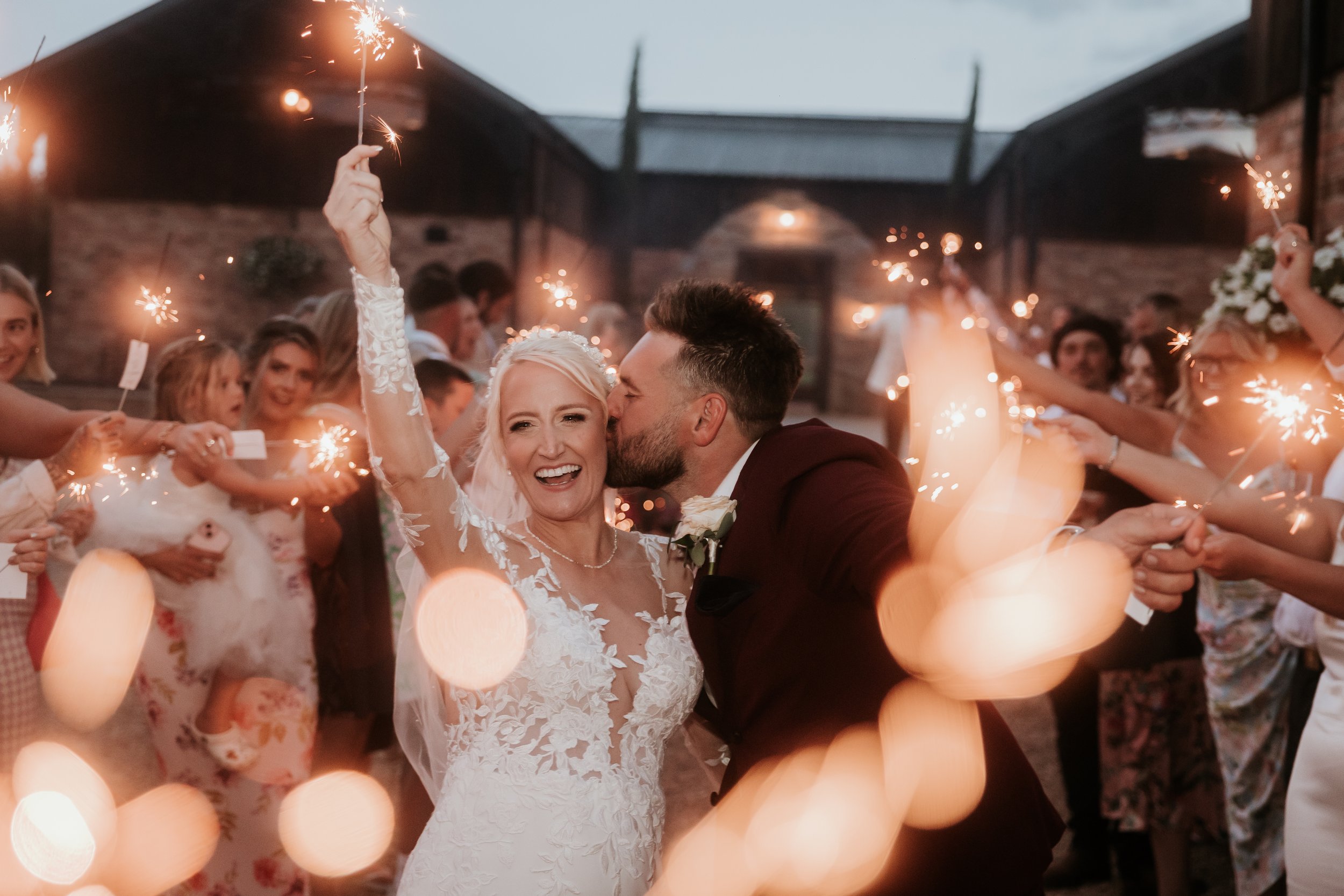 Bride and groom smiling, surrounded by guests holding sparklers at outdoor wedding celebration.