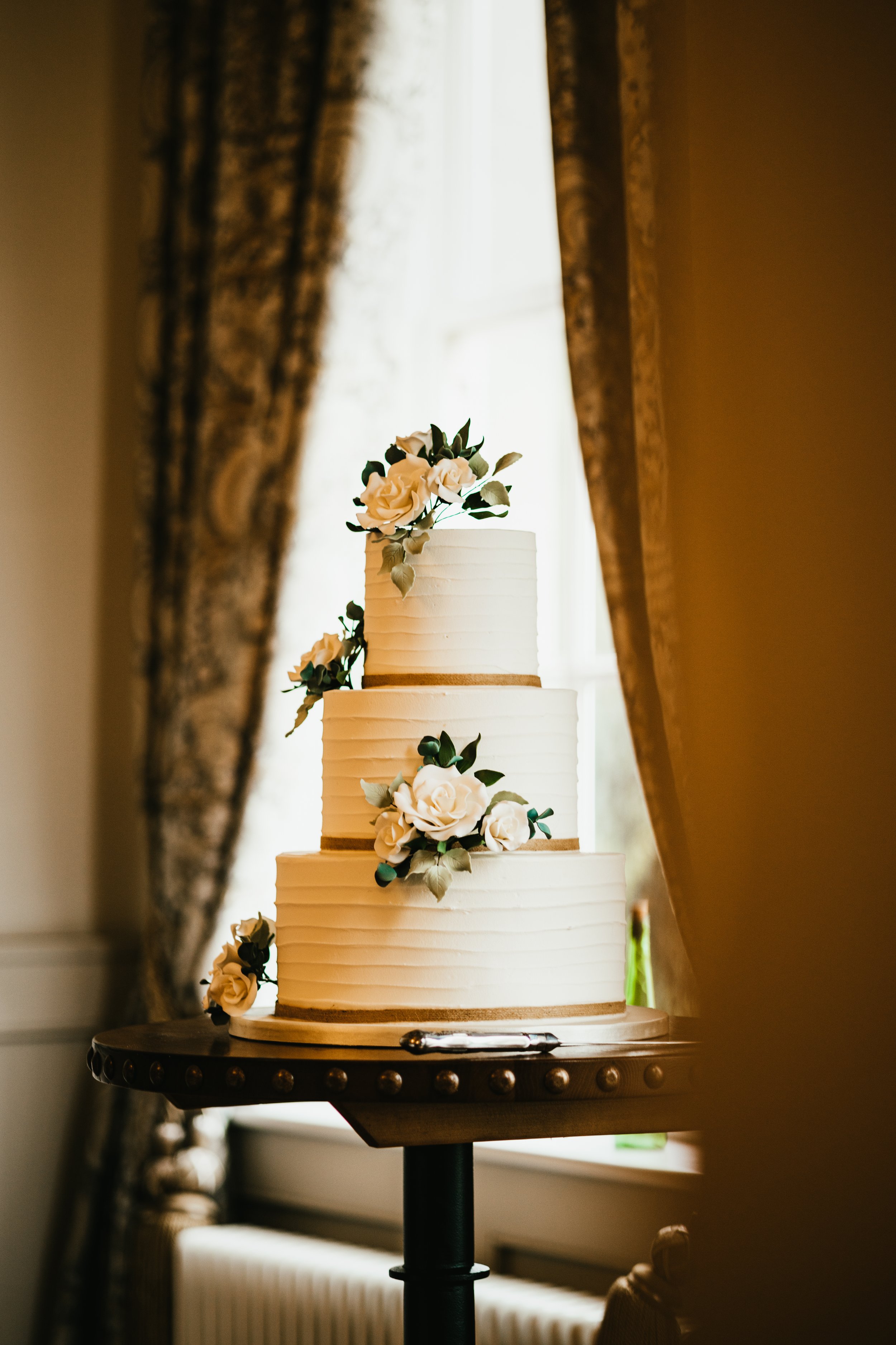 Three-tier white wedding cake with floral decorations on a wooden table, in a room with elegant curtains.
