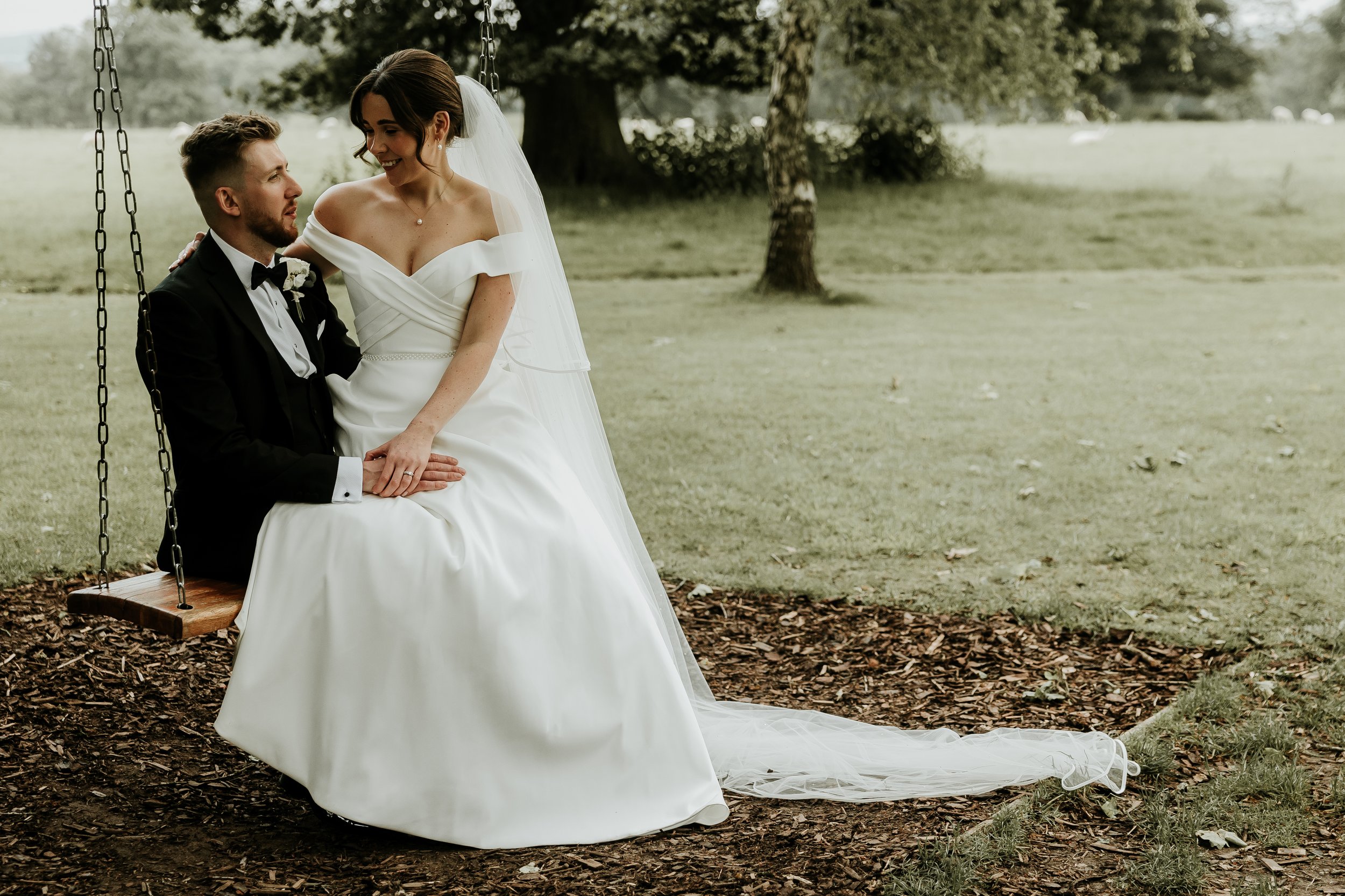 Bride in white dress and groom in tuxedo sitting on a wooden swing in a park, smiling at each other.