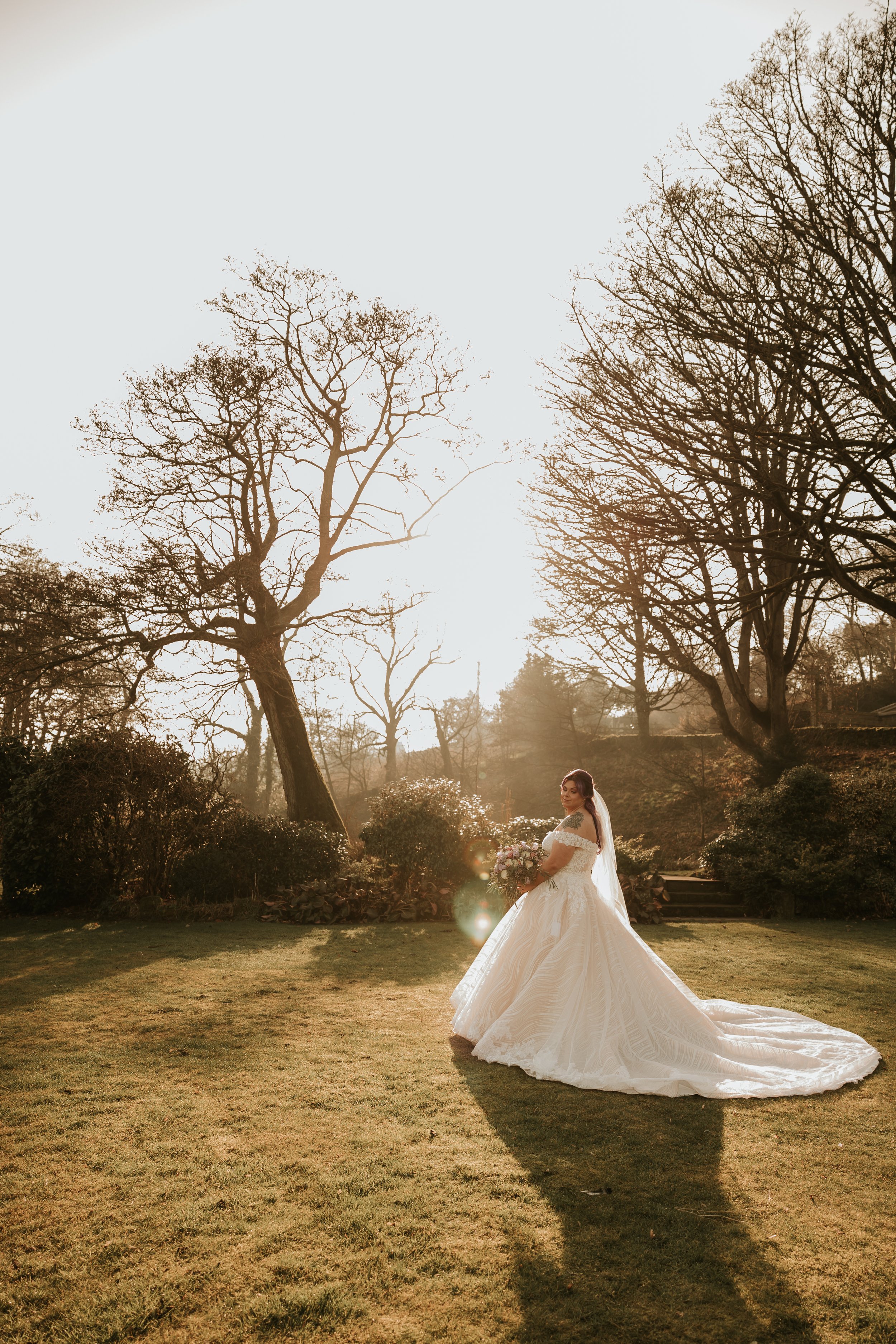 Bride in elegant gown holding bouquet in sunlit garden with leafless trees and green grass.