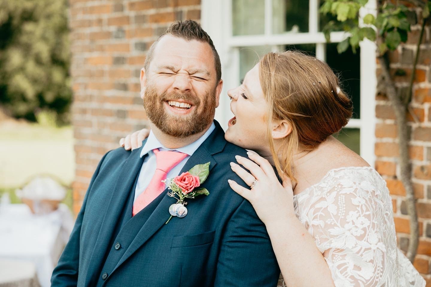A joyful couple, with a bearded man in a suit and pink tie smiling while a woman in a white lace dress playfully leans on his shoulder, outdoors near a brick wall with greenery.