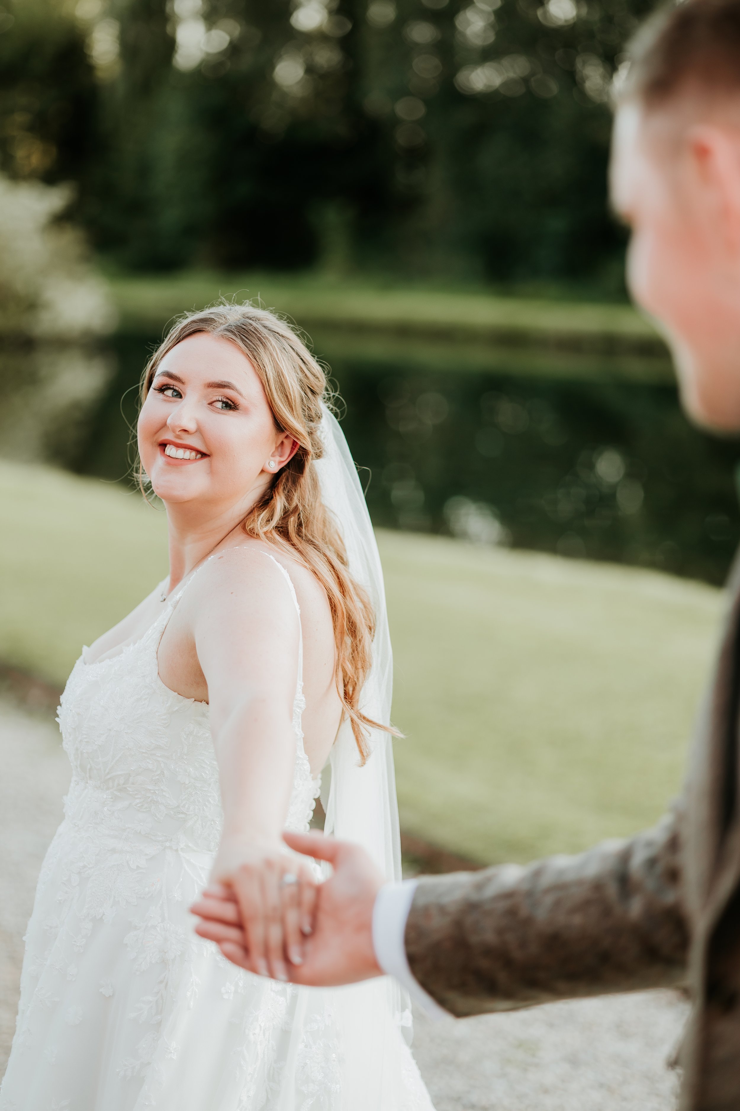 Bride smiling, holding hands with a man in a suit outdoors, near a pond.