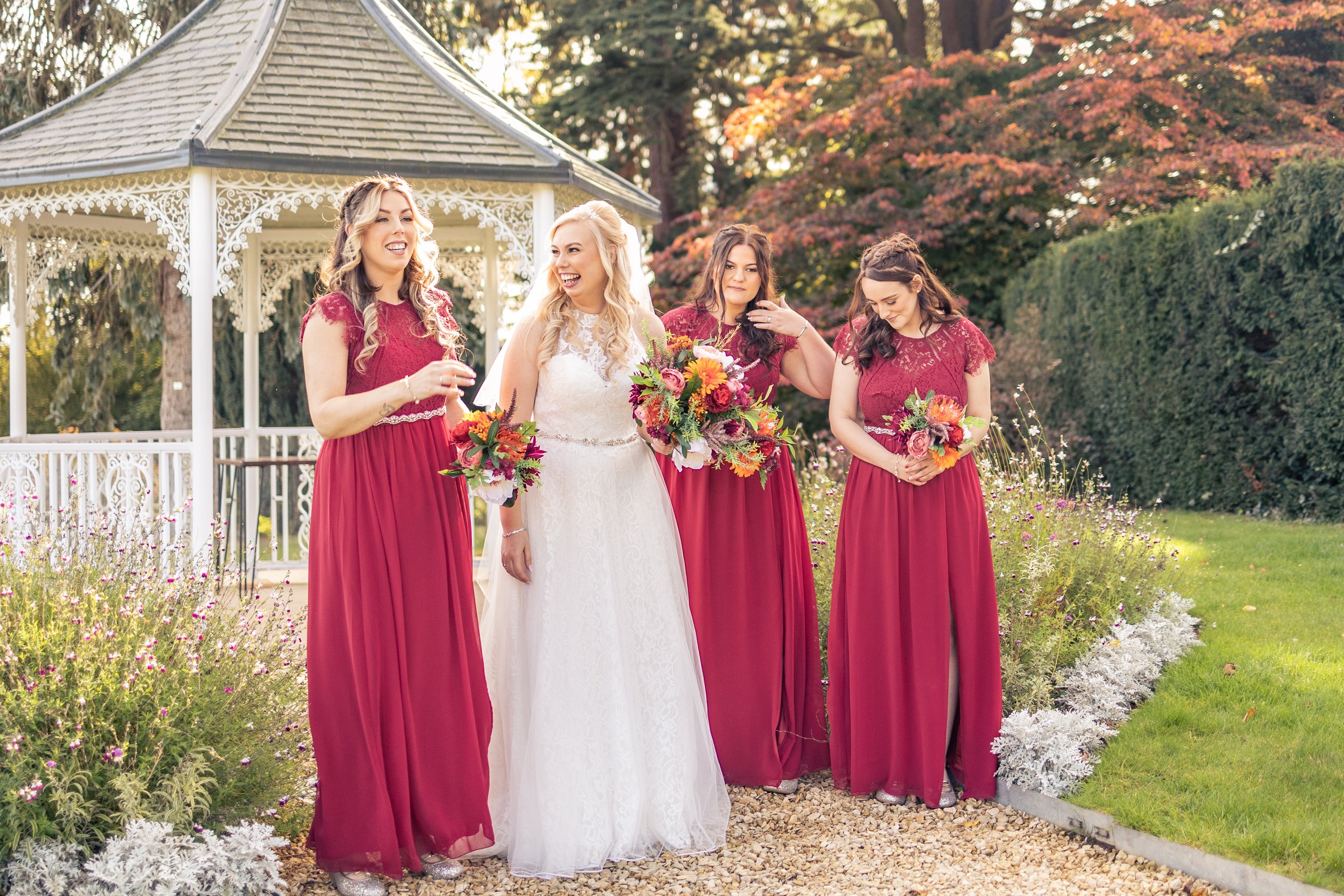 Bride with bridesmaids in red dresses standing by a gazebo in a garden setting.