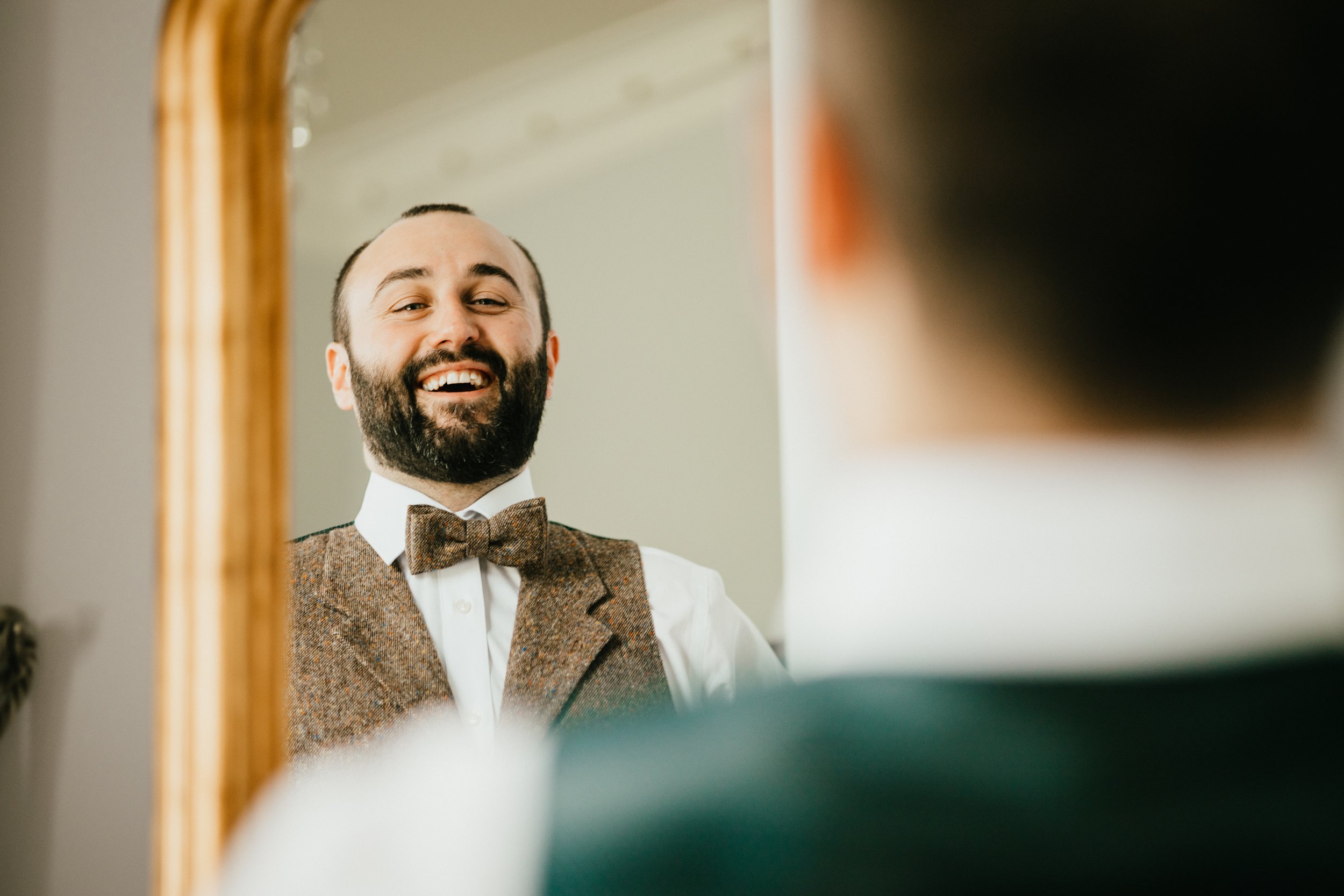 Man with a beard, wearing a tweed vest and bowtie, smiling in a mirror.