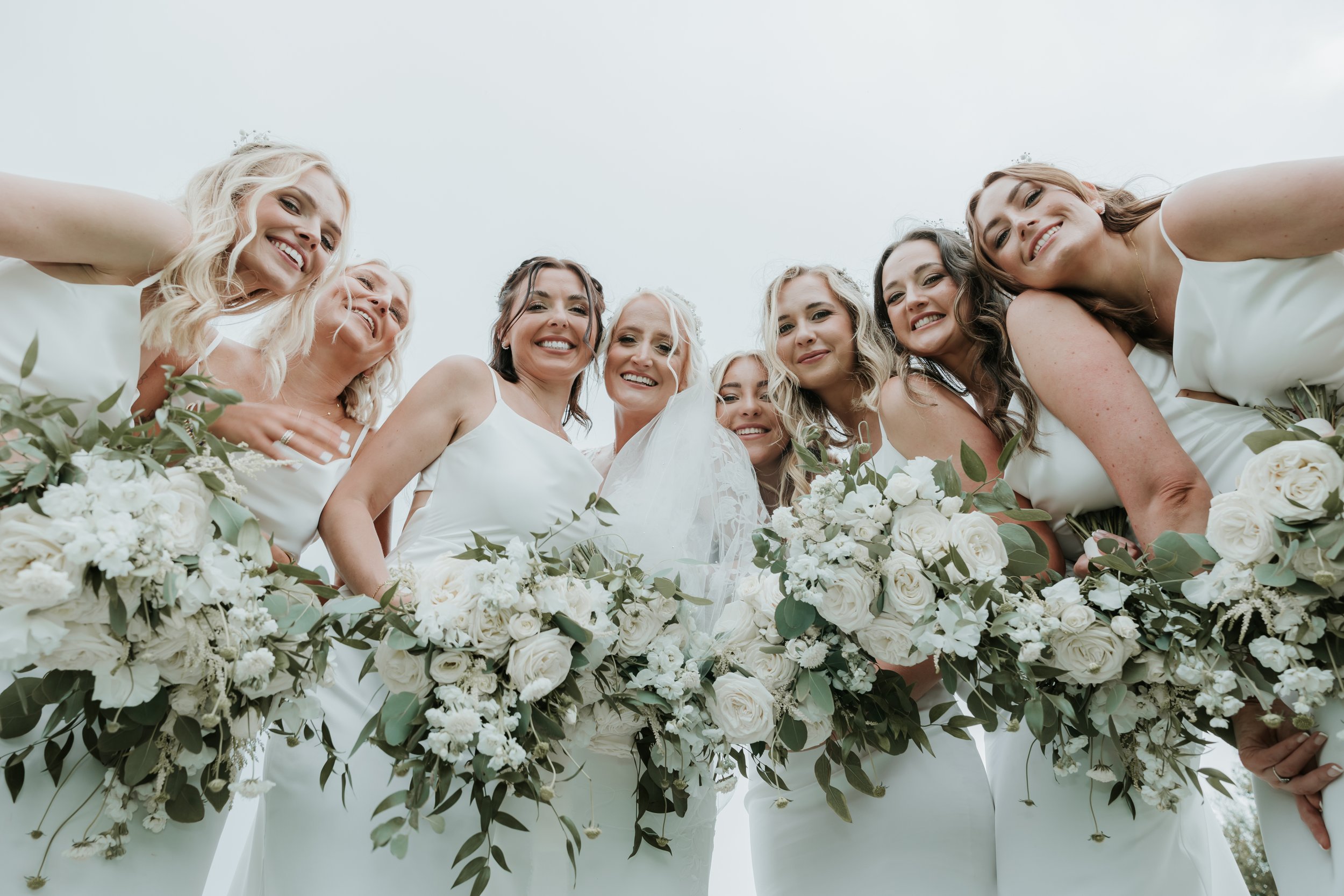 Bride and bridesmaids smiling and holding white floral bouquets, wearing white dresses.