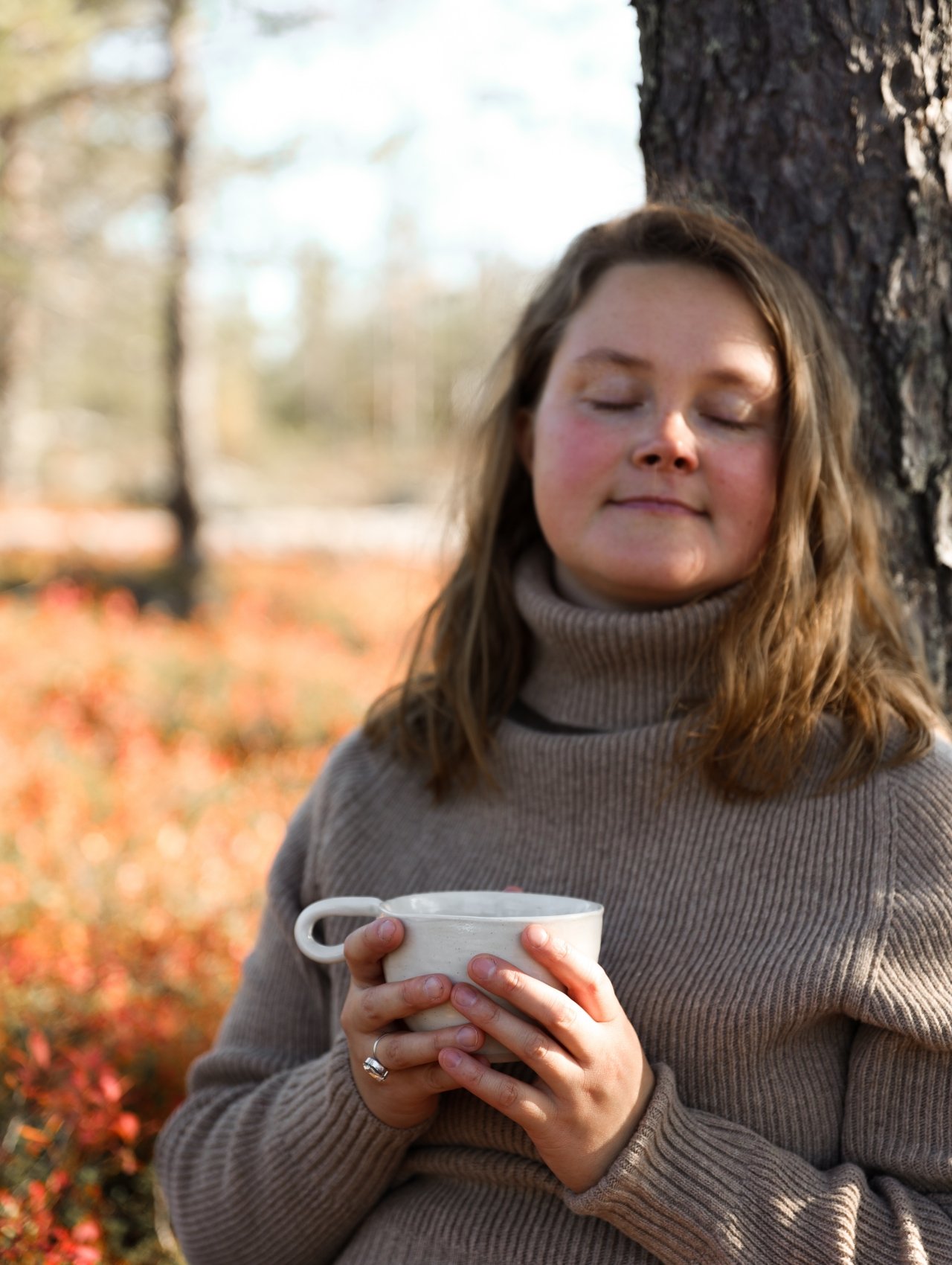 Nuori nainen seisoo puun vieressä ulkona syksyllä, hänellä on lämmin beige neule ja hän pitää kädessään kuppia, jossa on kuumaa juomaa, kasvoilla rauhallinen ilme.