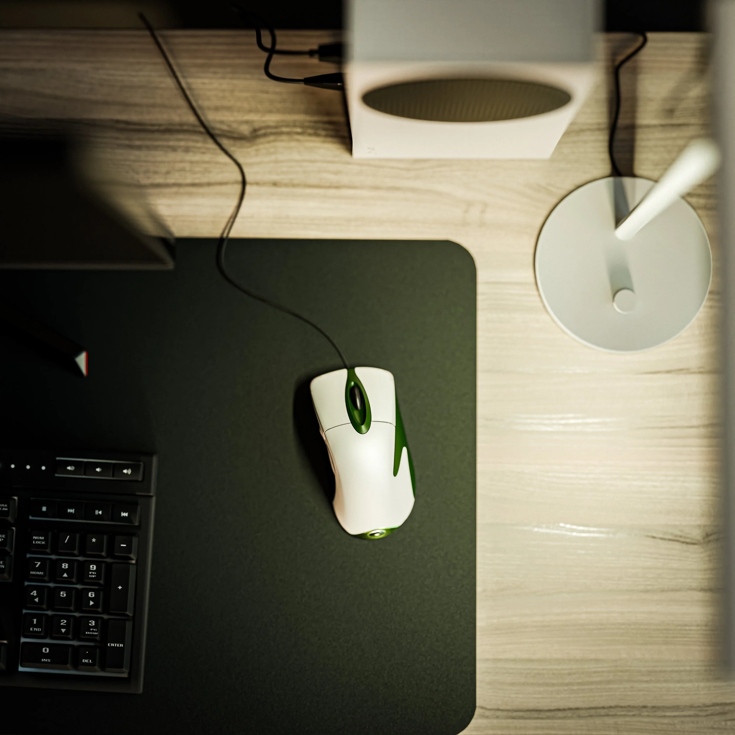 Souris d'ordinateur blanche et verte sur un tapis noir, à côté d'un clavier partiel, sur un bureau en bois, avec une lampe et une enceinte Bluetooth.