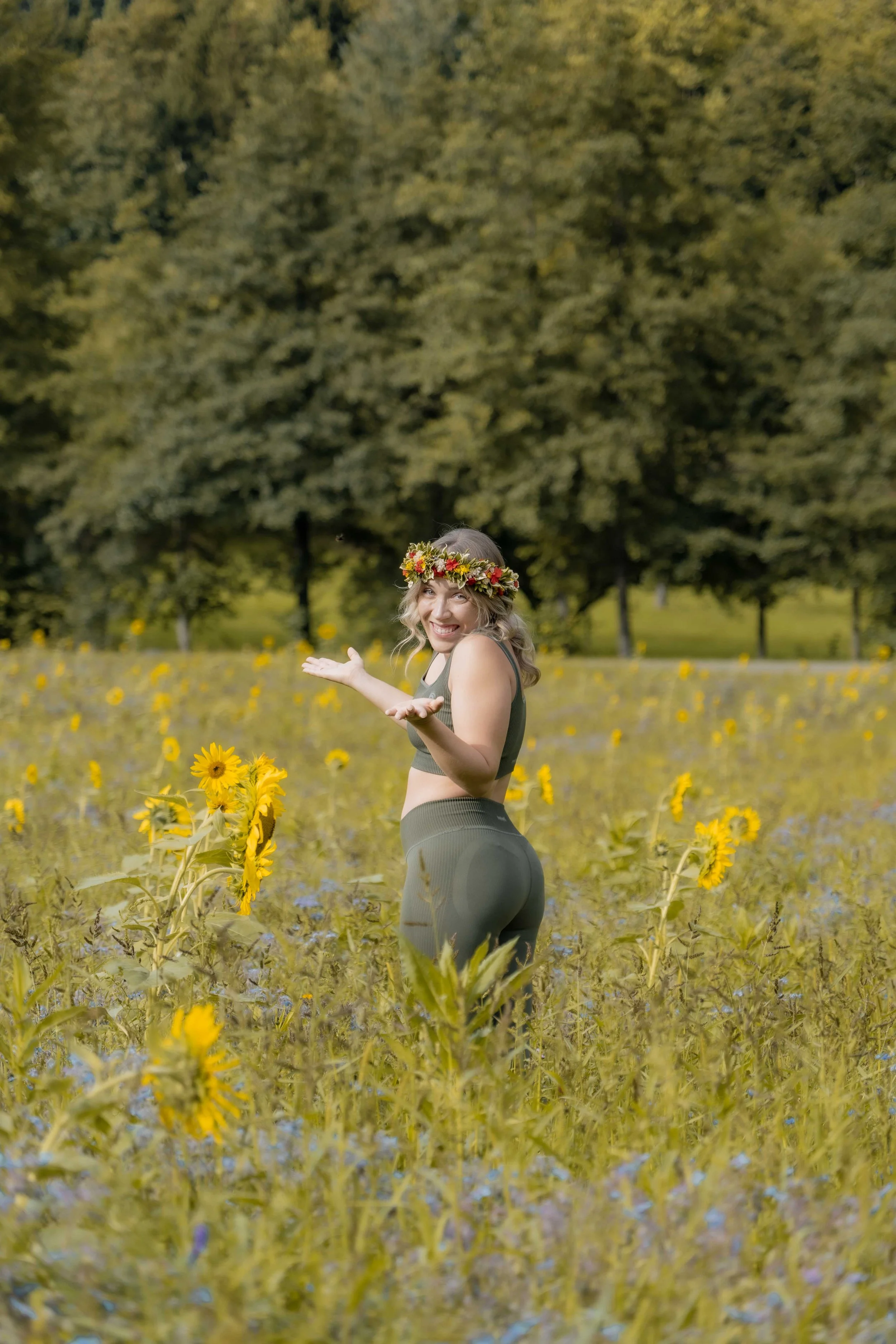 Junge Frau im Sonnenblumenfeld mit Blumenkranz im Haar, lächelnd und mit offenen Armen. Hintergrund aus Bäumen.