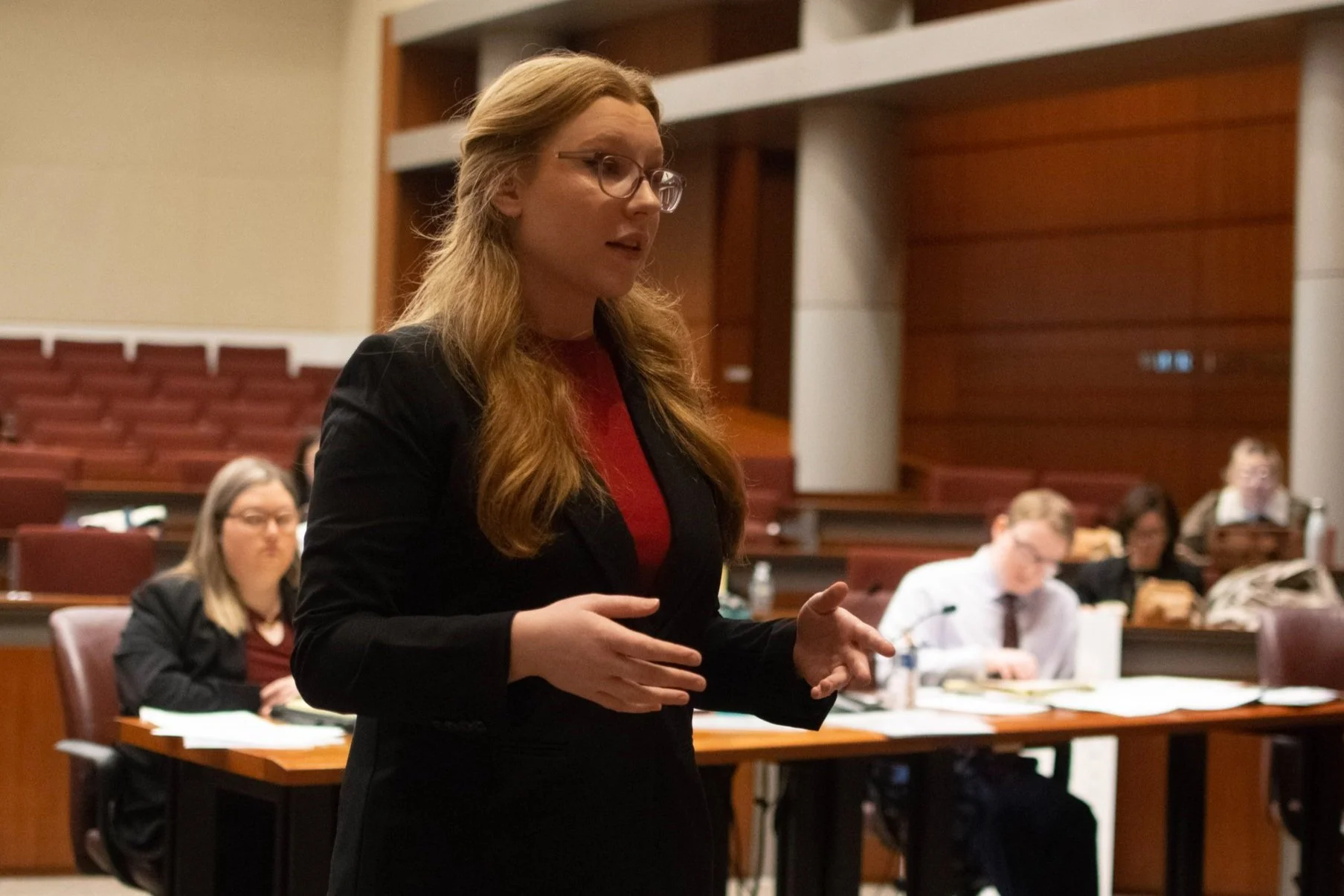 A woman with long blonde hair, glasses, and a black blazer speaking in a formal setting with other people seated at tables and taking notes, in a room with wooden panels.