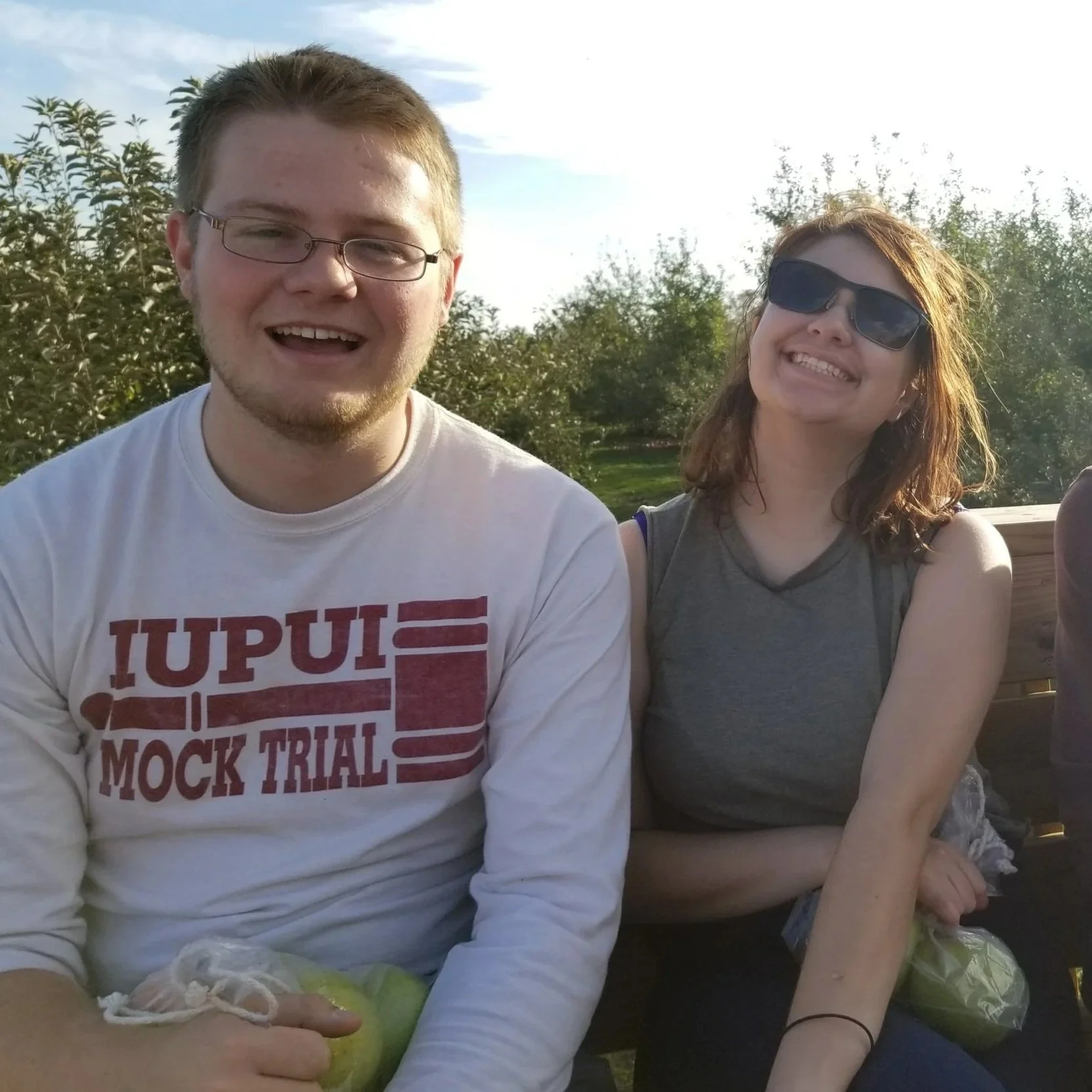 Two friends sitting outside on a sunny day, smiling. The person on the left has glasses and a white shirt with red text, the person on the right has glasses and a sleeveless gray shirt. There are trees and a bright sky in the background.