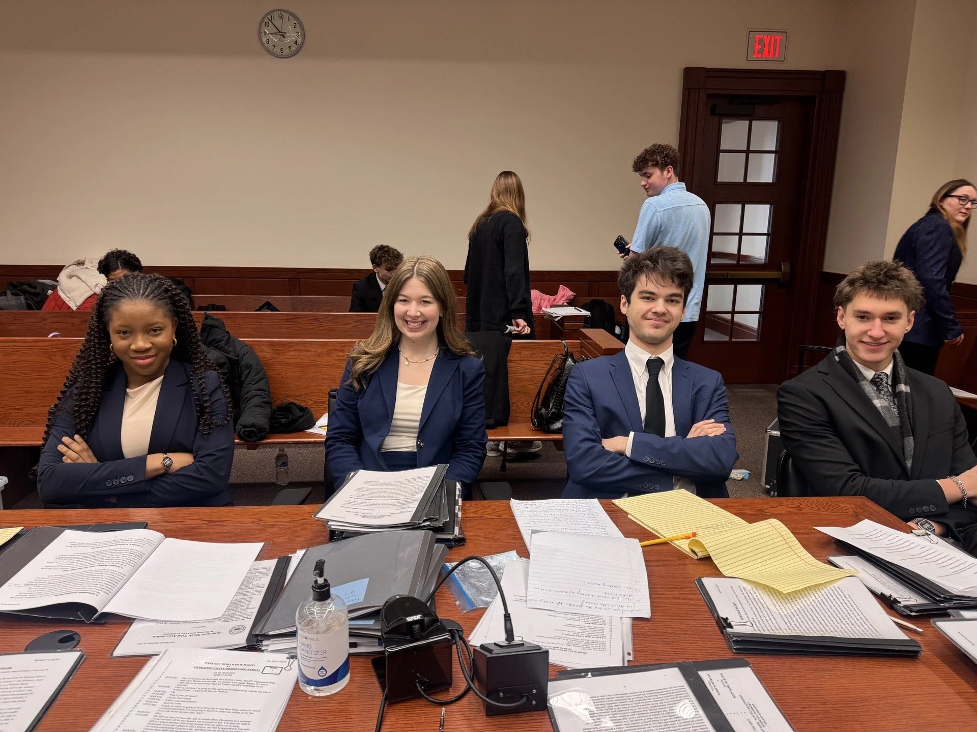 Group of people in a professional setting, with three individuals in the foreground smiling and seated at a desk, and several others in the background in a meeting or conference room.
