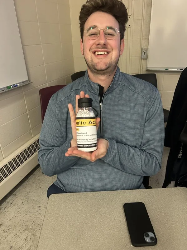 A man with glasses smiling and sitting at a table, holding a bottle labeled 'Salicylic Acid' with both hands.