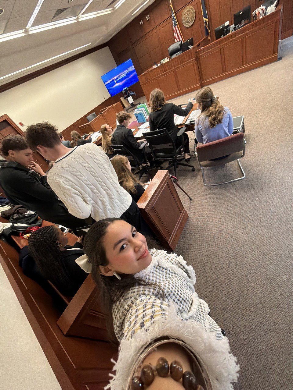 A group of people in a courtroom, with a woman taking a selfie in the foreground. The courtroom has wood paneling, American flags, and a large screen at the front.