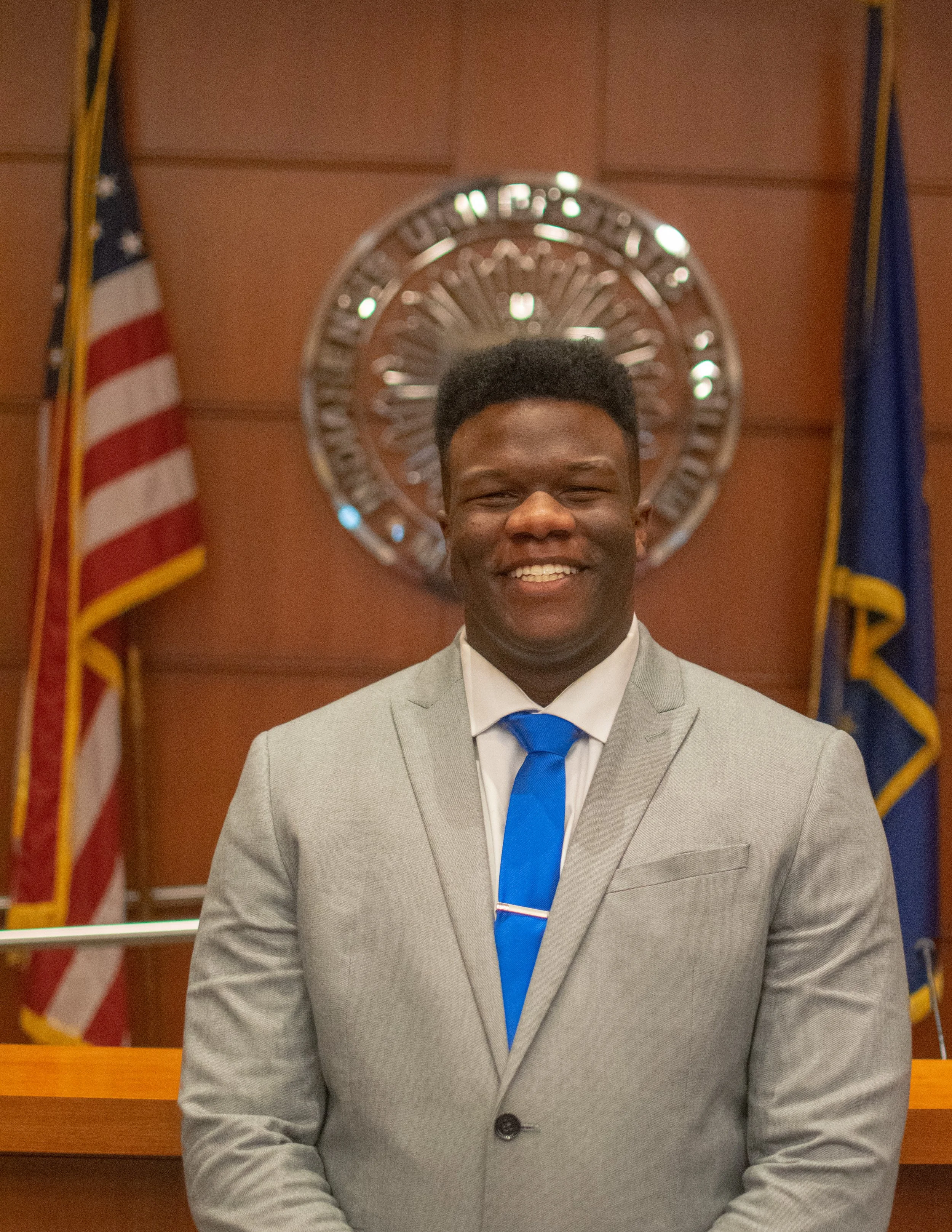 Portrait of a young man in a gray suit, light blue tie, smiling in front of an American flag and a government emblem.