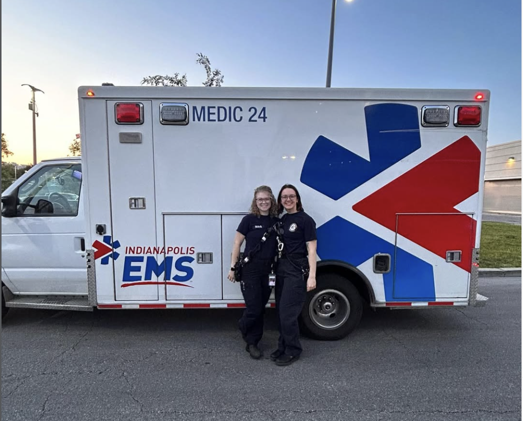 Two female emergency medical service providers standing in front of an ambulance with Indianapolis EMS logo, smiling and posing outdoors during sunset.