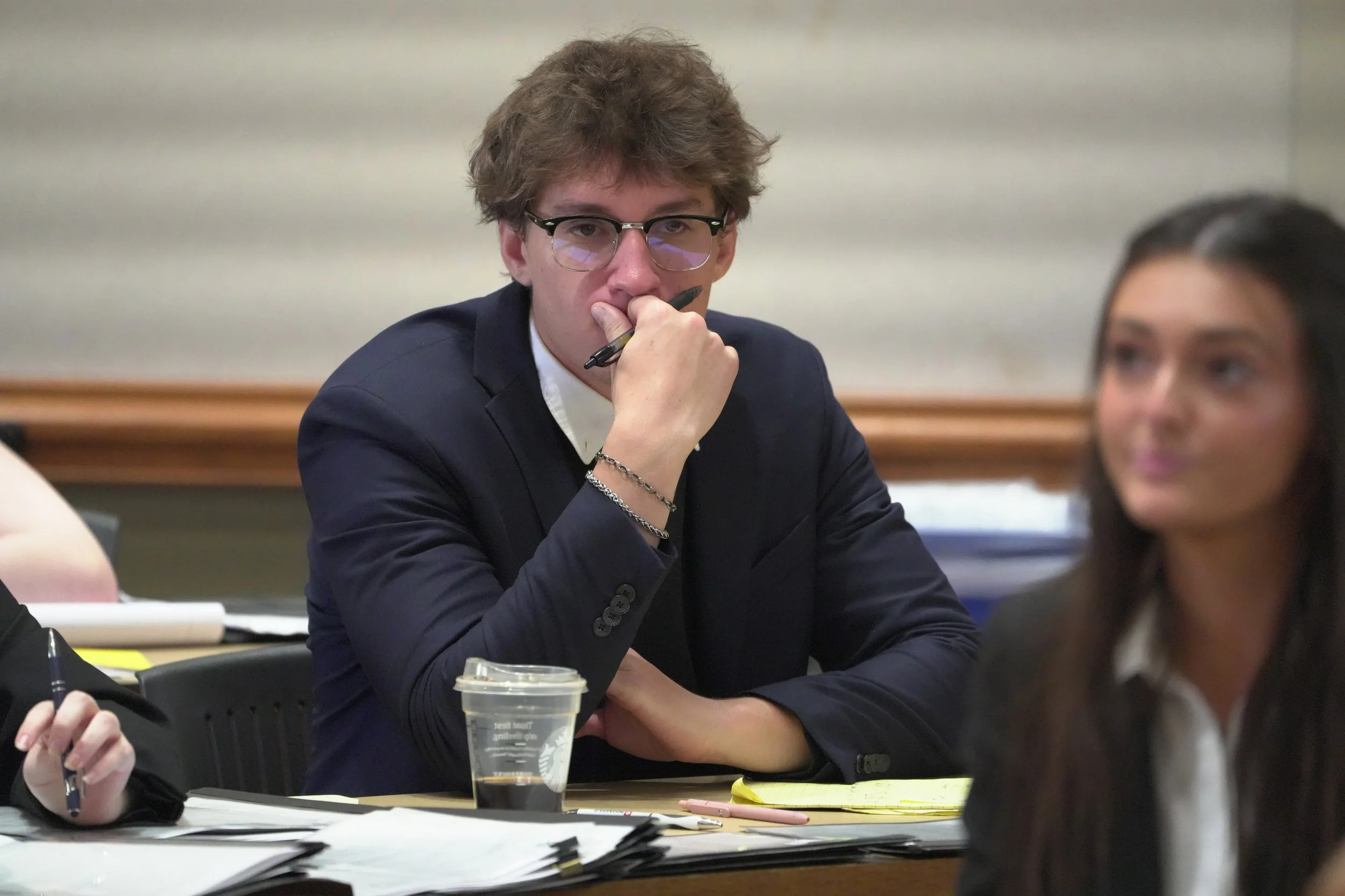 A young man with glasses and wavy hair, dressed in a blazer, sitting at a table with papers, a notebook, and a cup of coffee, appears to be in a serious discussion or meeting.