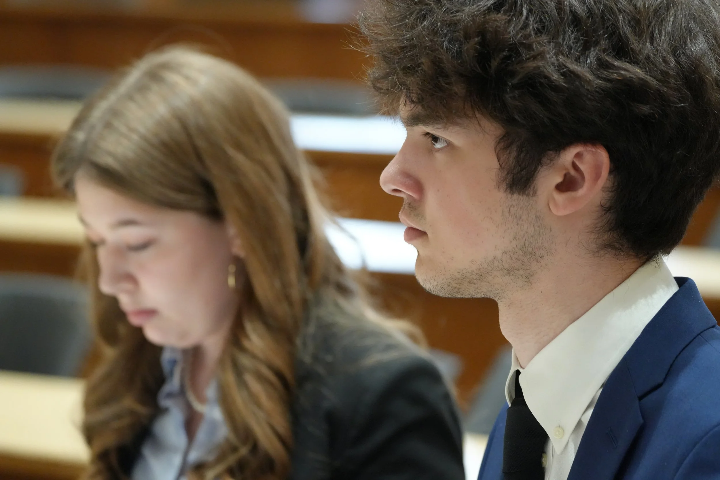 Close-up side profile of a young man with curly dark hair wearing a navy blue suit and white shirt, sitting in a classroom setting with other students.