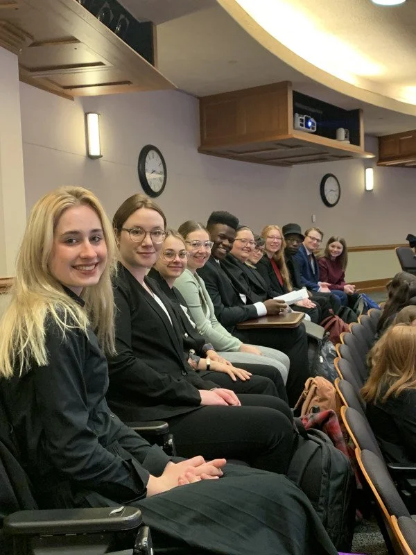 Group of young adults seated in a conference room, smiling at the camera. They are sitting in chairs along a wall with a clock and wall-mounted lights visible.