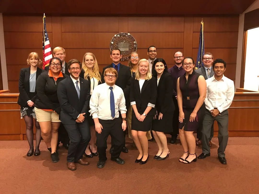 Group of diverse young adults dressed in business attire, standing in a courtroom or formal setting with wooden paneling, American flags, and a official seal in the background.