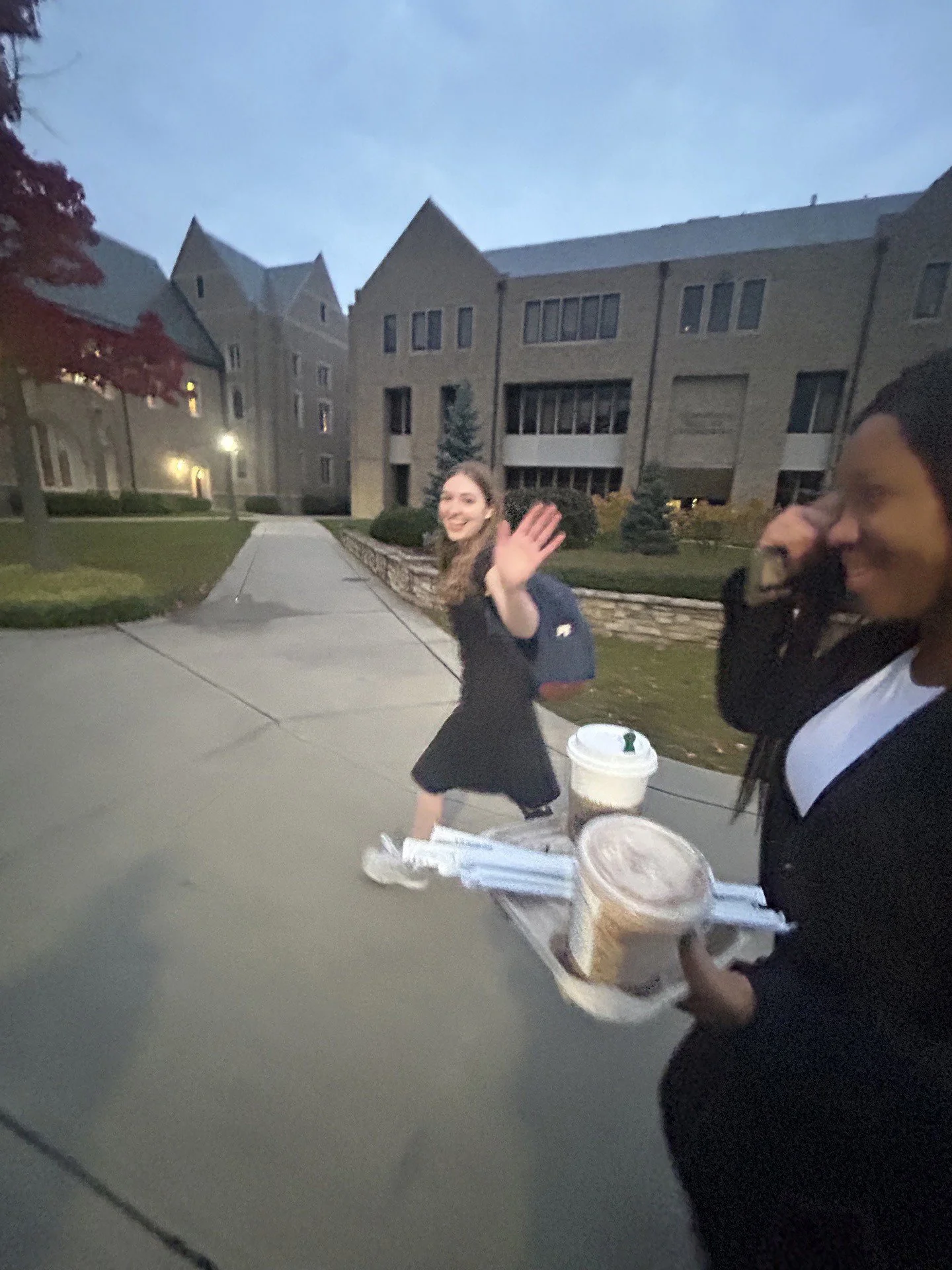 A woman in a black dress waving and smiling, another woman holding drinks and food in a takeout tray with a large building in the background.