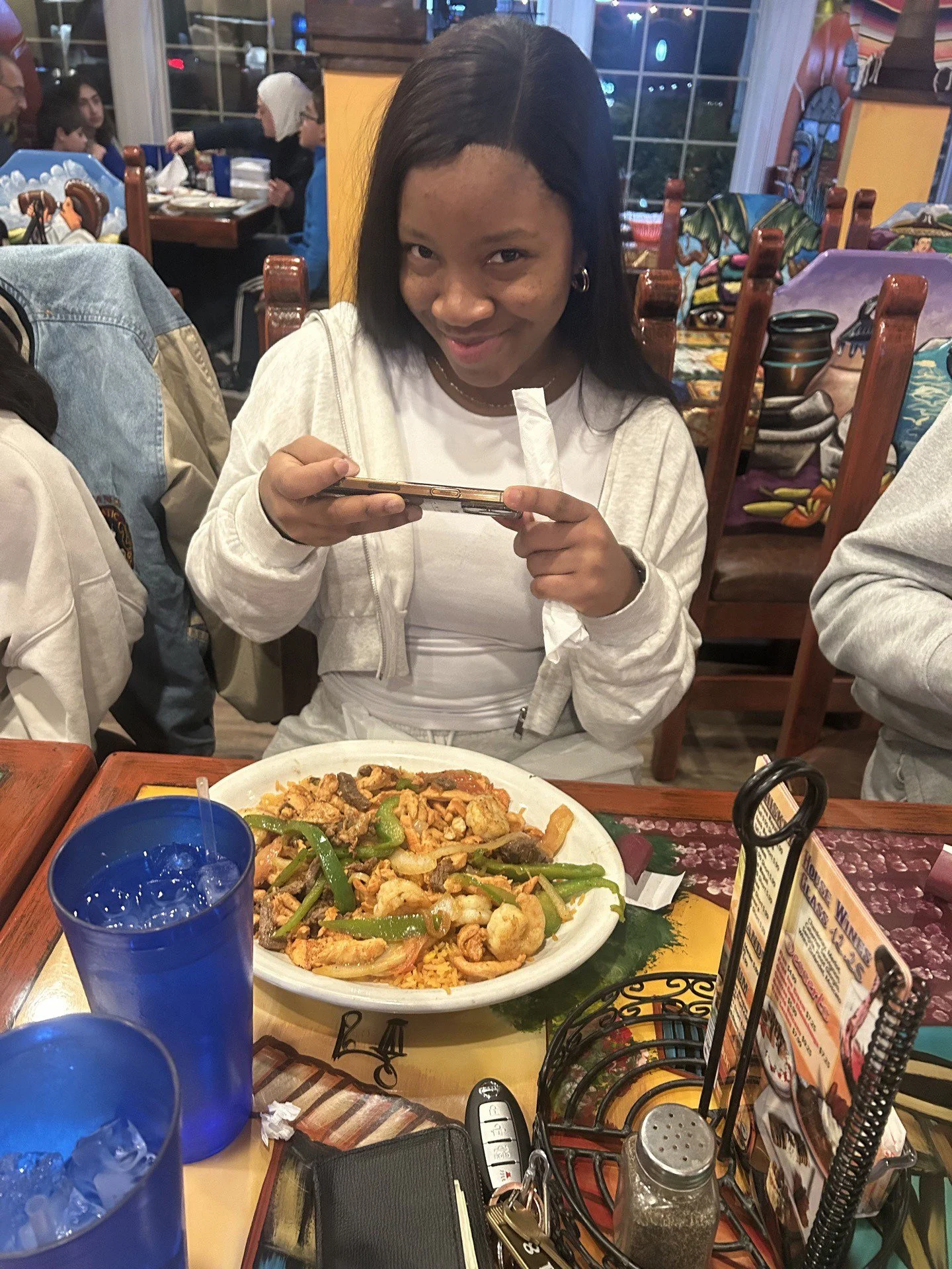 A woman with black hair smiling and looking at the camera, sitting at a restaurant table with a plate of shrimp and vegetables, a blue drink, and various restaurant items on the table.