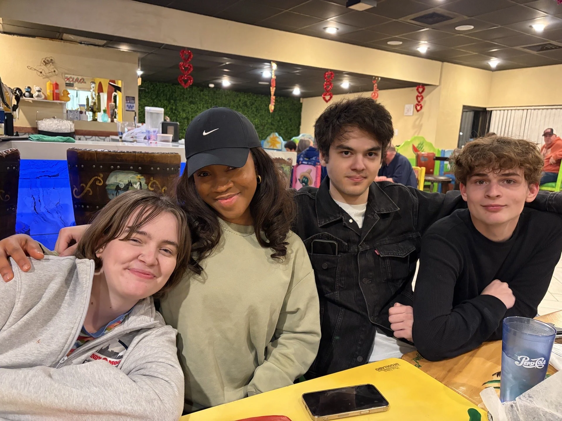 Four people sitting at a table in a colorful restaurant or cafe, smiling and posing for a photo. The background features festive decorations, a bar area, and bright walls.