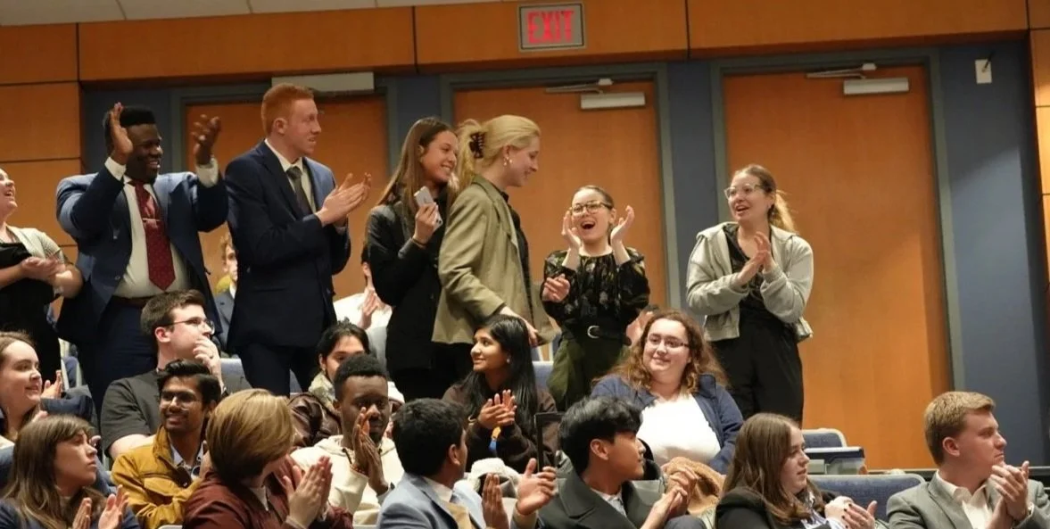 Group of people clapping and smiling in an auditorium, some standing and some seated, with a wooden-paneled wall and exit sign in the background.